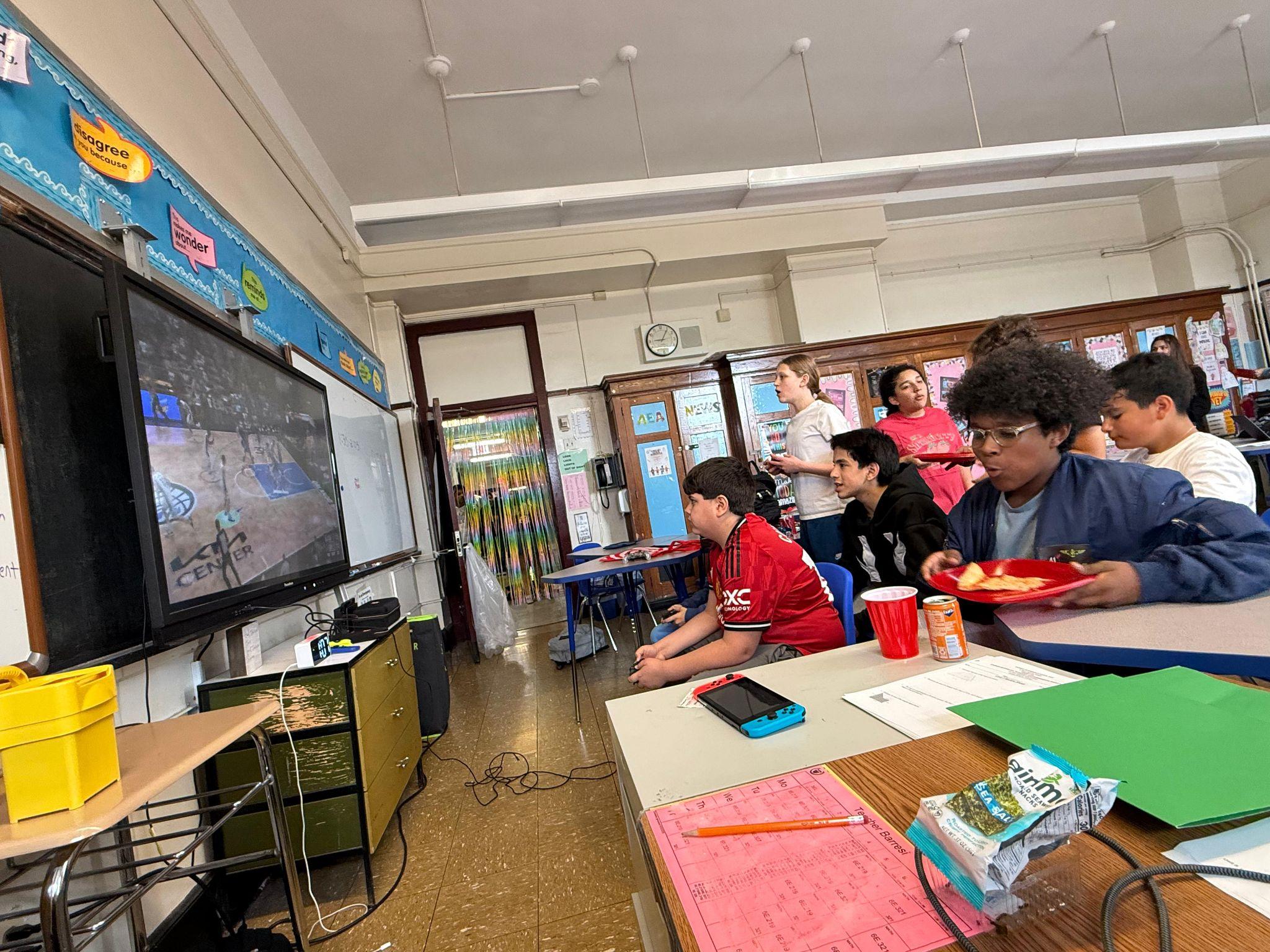 Students watching a game on a classroom TV with snacks