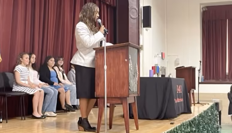 Woman speaks at podium during school ceremony with students seated on stage