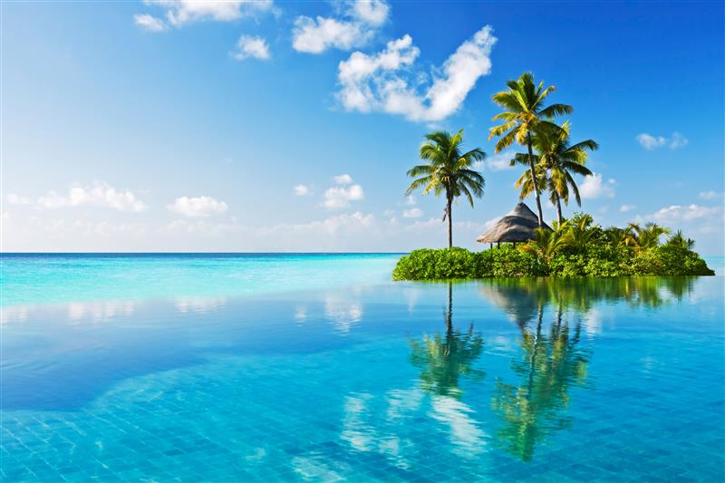 Small tropical island with palm trees and a hut reflected in calm blue ocean water under a blue sky.