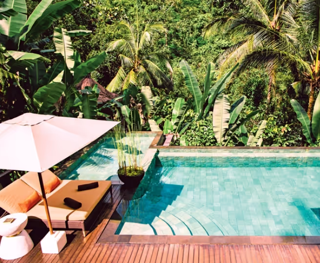 Outdoor pool with wooden deck, lounge chair, umbrella, and tropical greenery in the background.