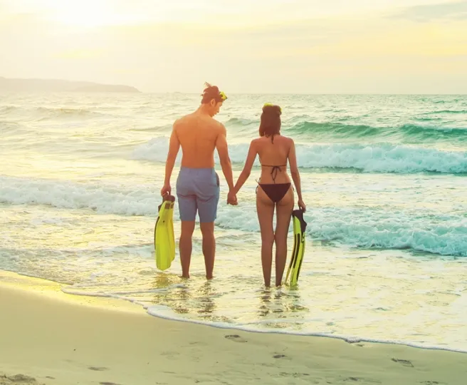 Couple holding hands at the beach shore with snorkeling fins, facing the ocean waves at sunset.