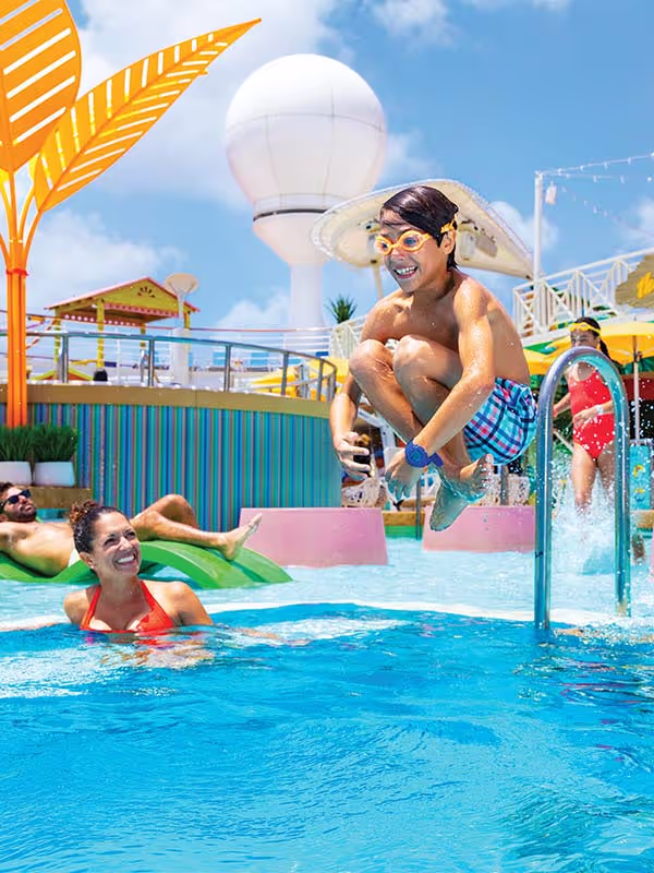 A boy wearing goggles jumps into a pool with a woman and man nearby on floaties on a sunny day.