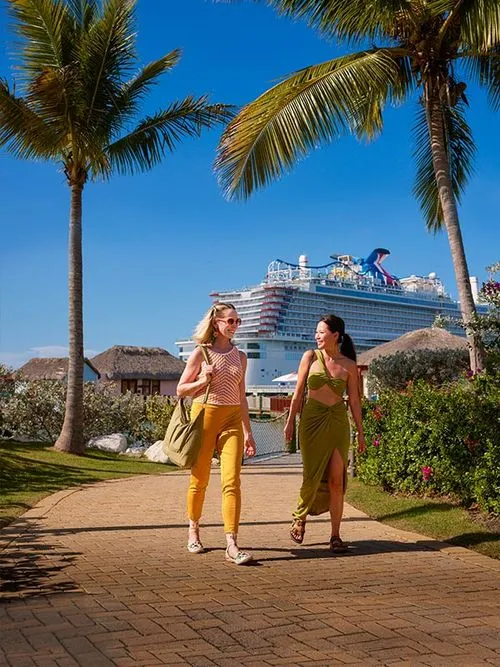 Two women walking on a path between palm trees with a large cruise ship docked nearby.