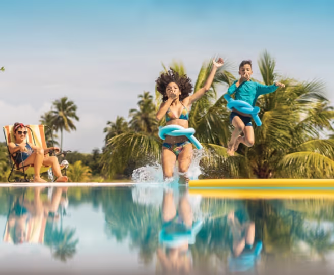 Two kids jumping into a pool with blue float belts, woman watching while reclining on a chair nearby.
