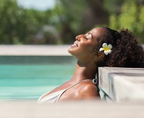 Woman with flower in hair relaxing with eyes closed, leaning against pool edge in sunny outdoor setting.