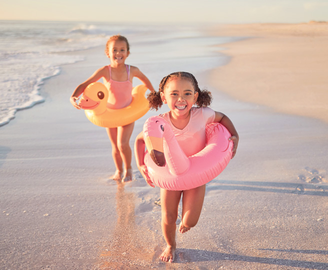 Two young girls running on the beach with inflatable swim rings shaped like a pink flamingo and a yellow duck.