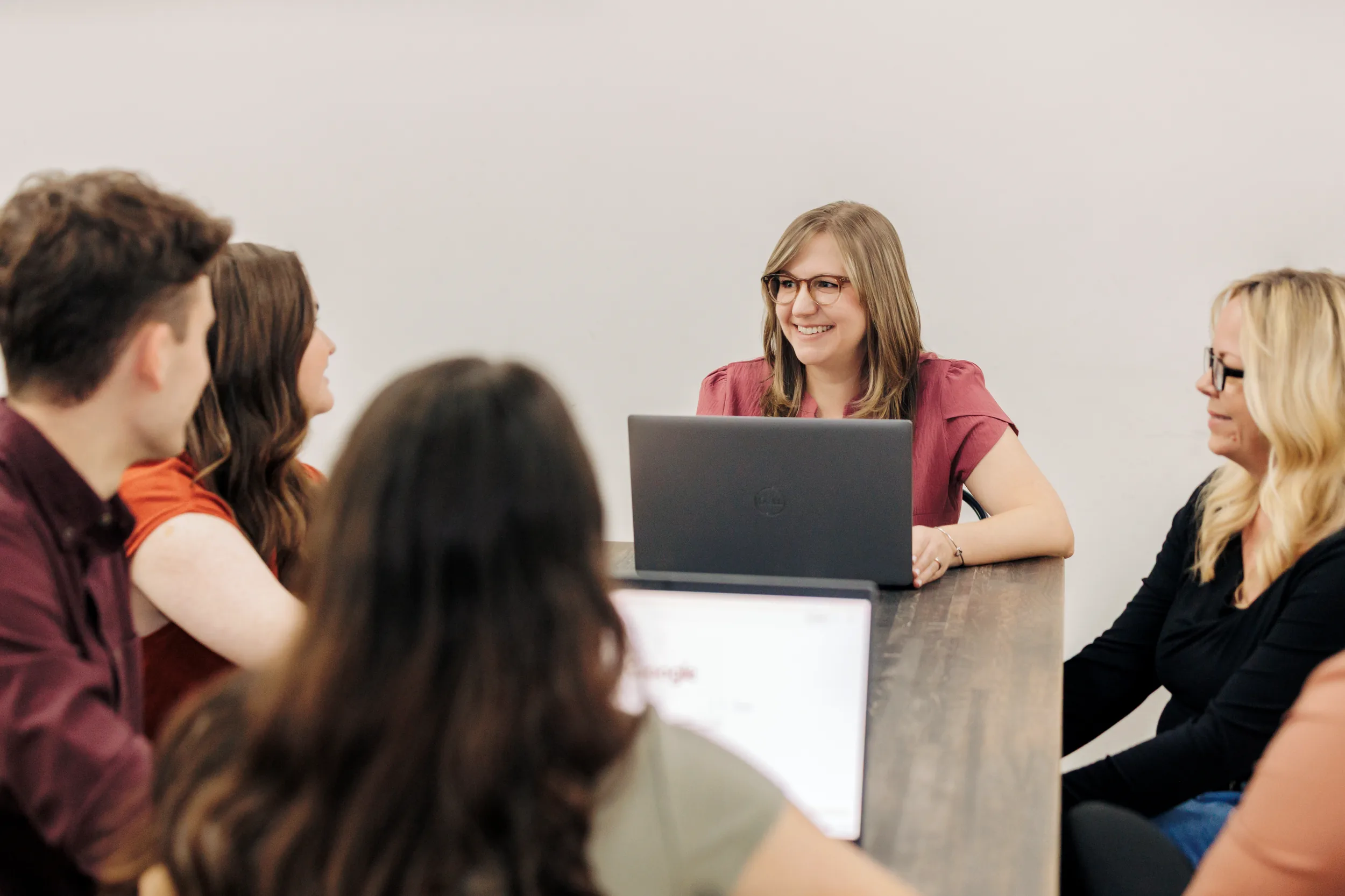 Group of five people sitting around a wooden table in a meeting room, two with laptops open, engaged in conversation, with a smiling woman wearing glasses and a red shirt at the head of the table.