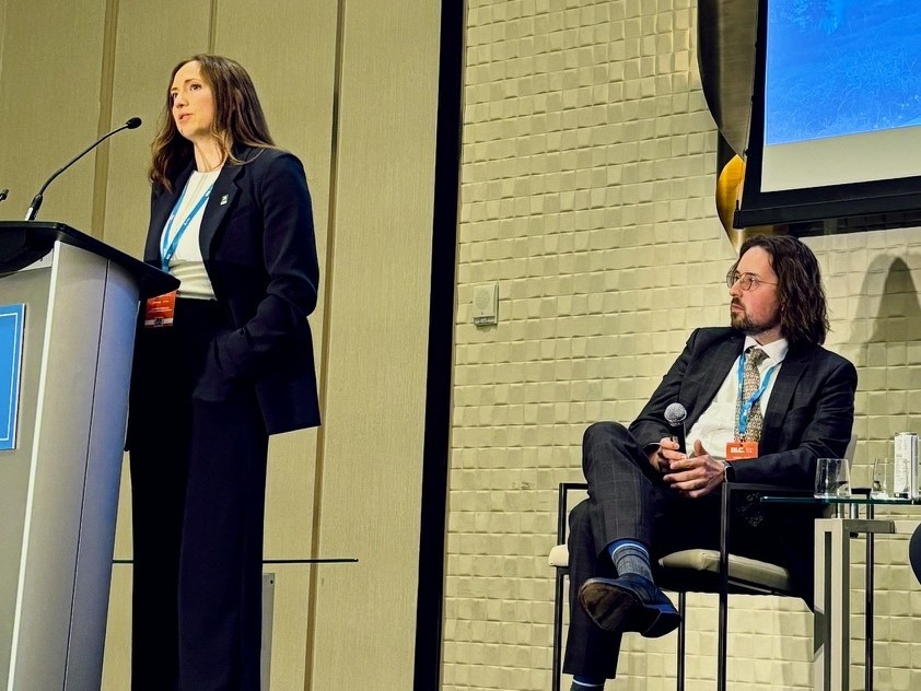 Ashley Sarauer speaking at a podium on a conference stage, with a fellow panelist seated beside her