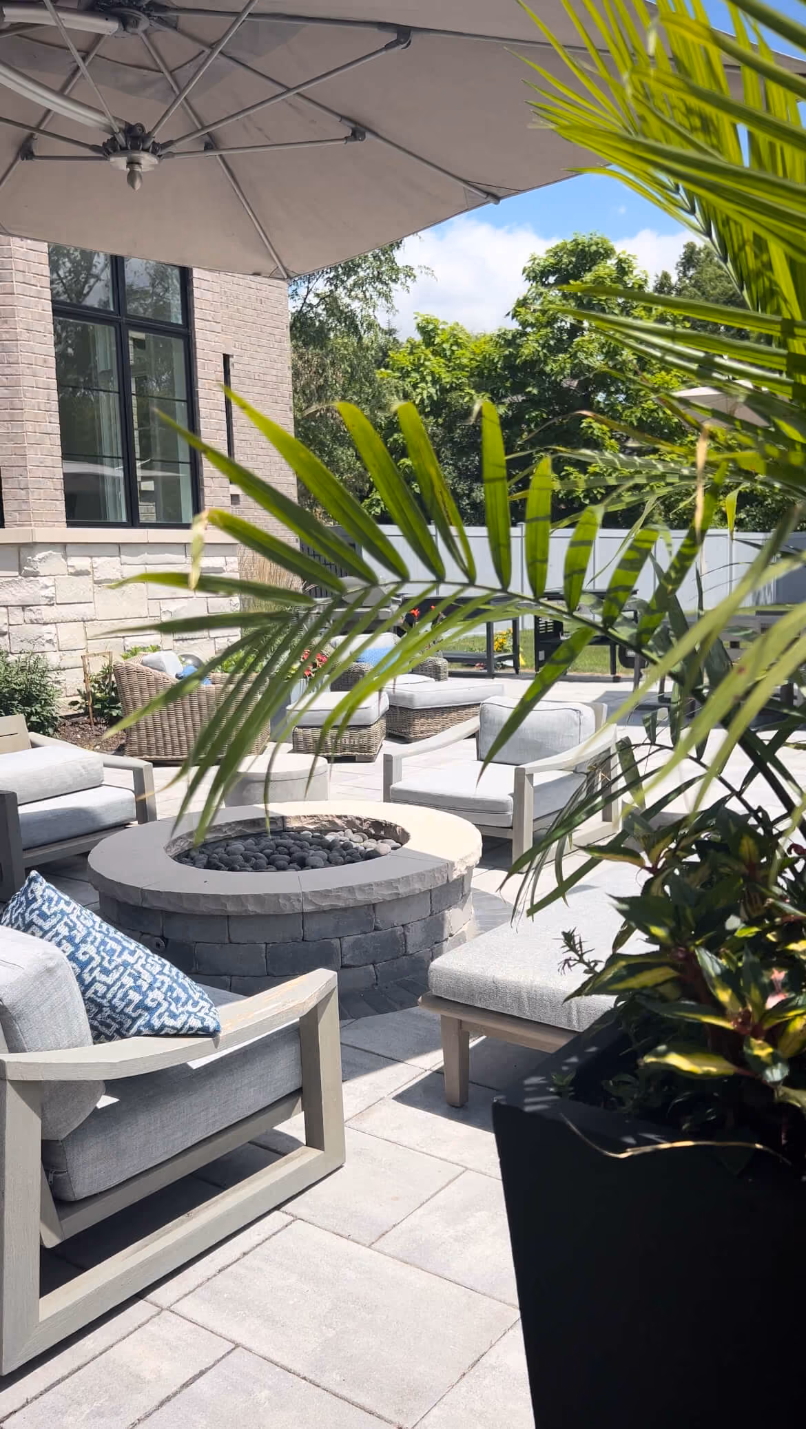 Modern backyard patio with cushioned chairs, a circular stone fire pit, umbrella, and green plants.
