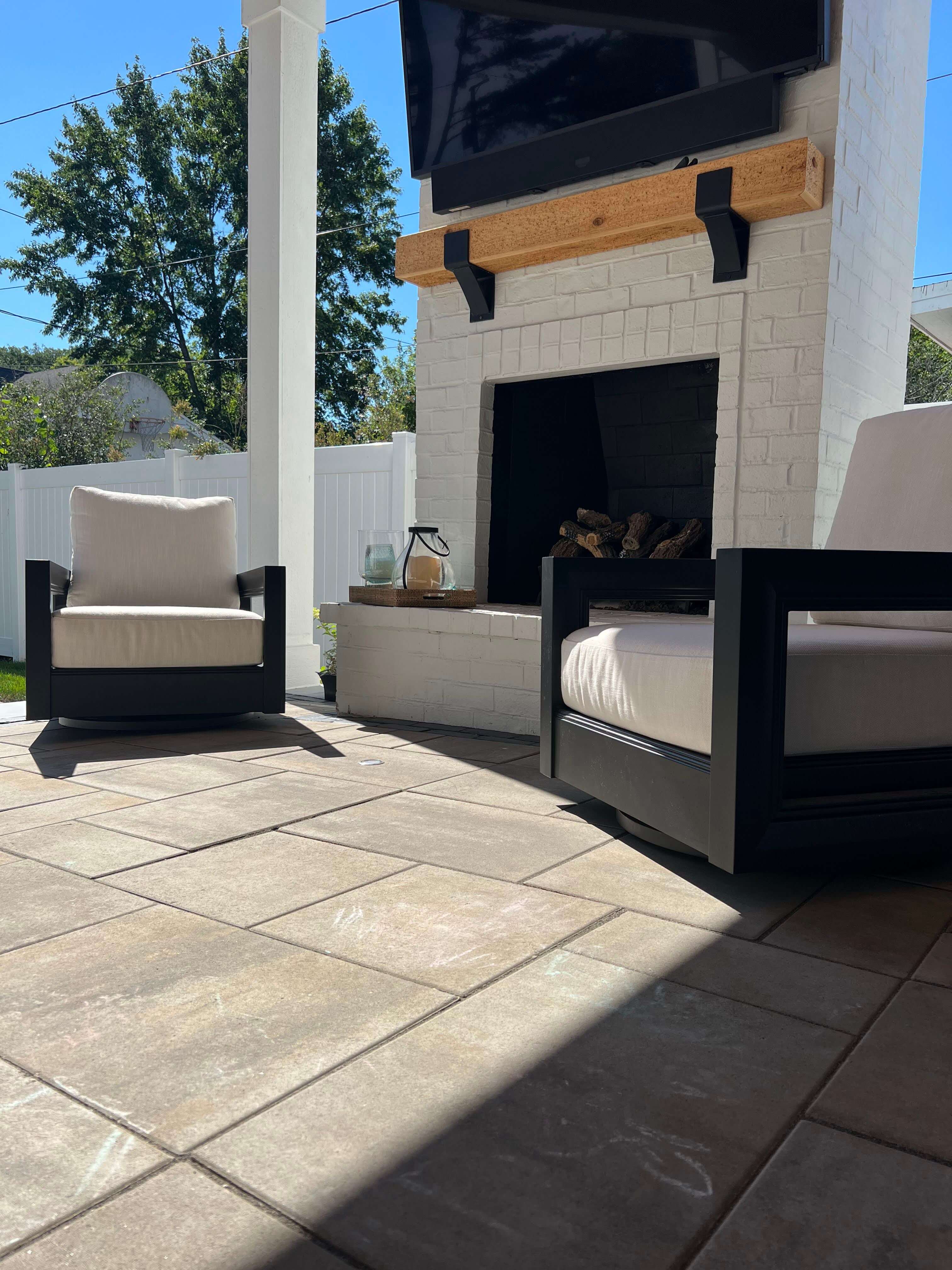 Outdoor patio with two cushioned black chairs, tiled floor, white brick fireplace, and TV mounted above.