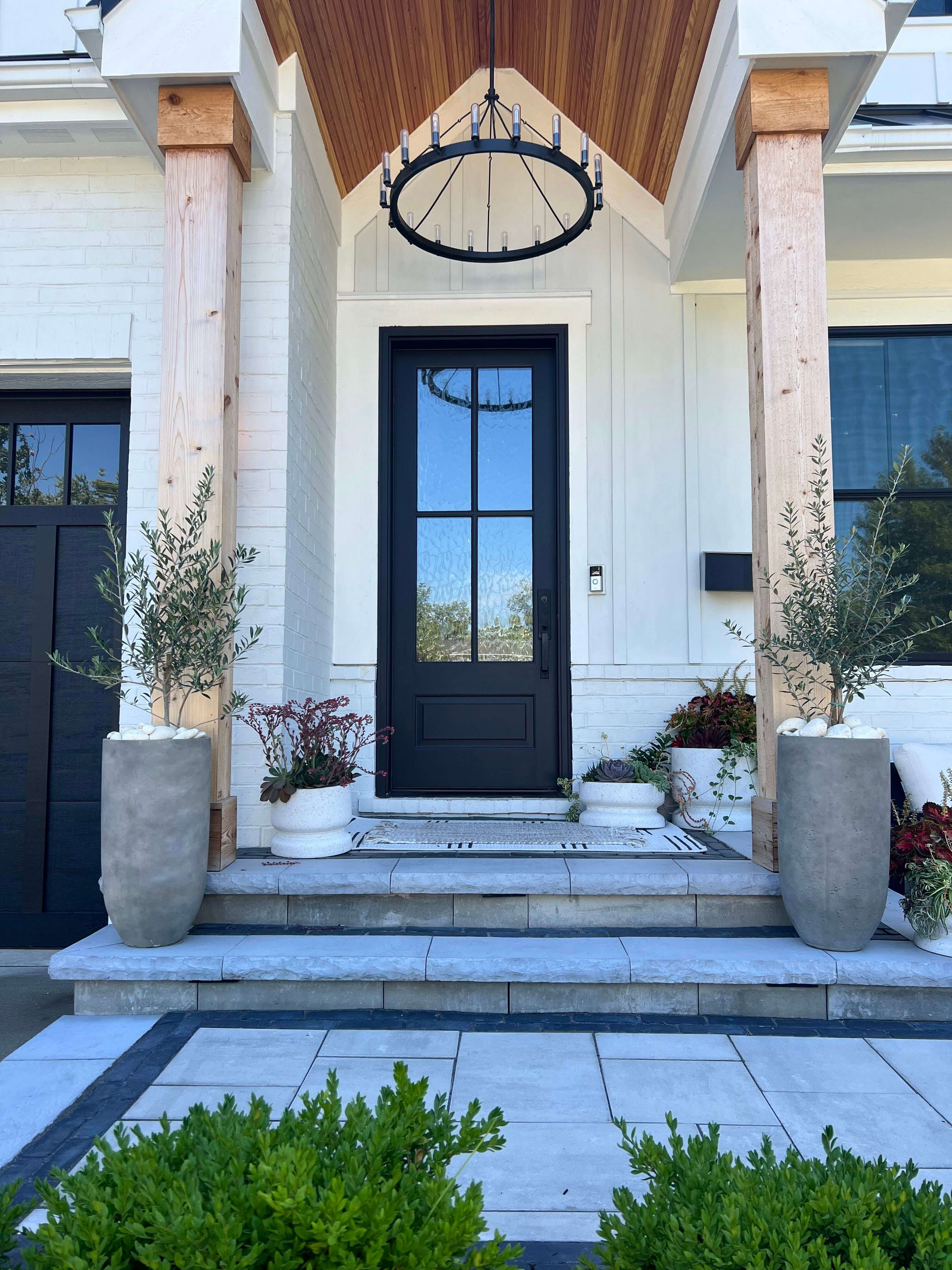 Front porch with black door, wooden pillars, hanging chandelier, stone steps, and potted plants.
