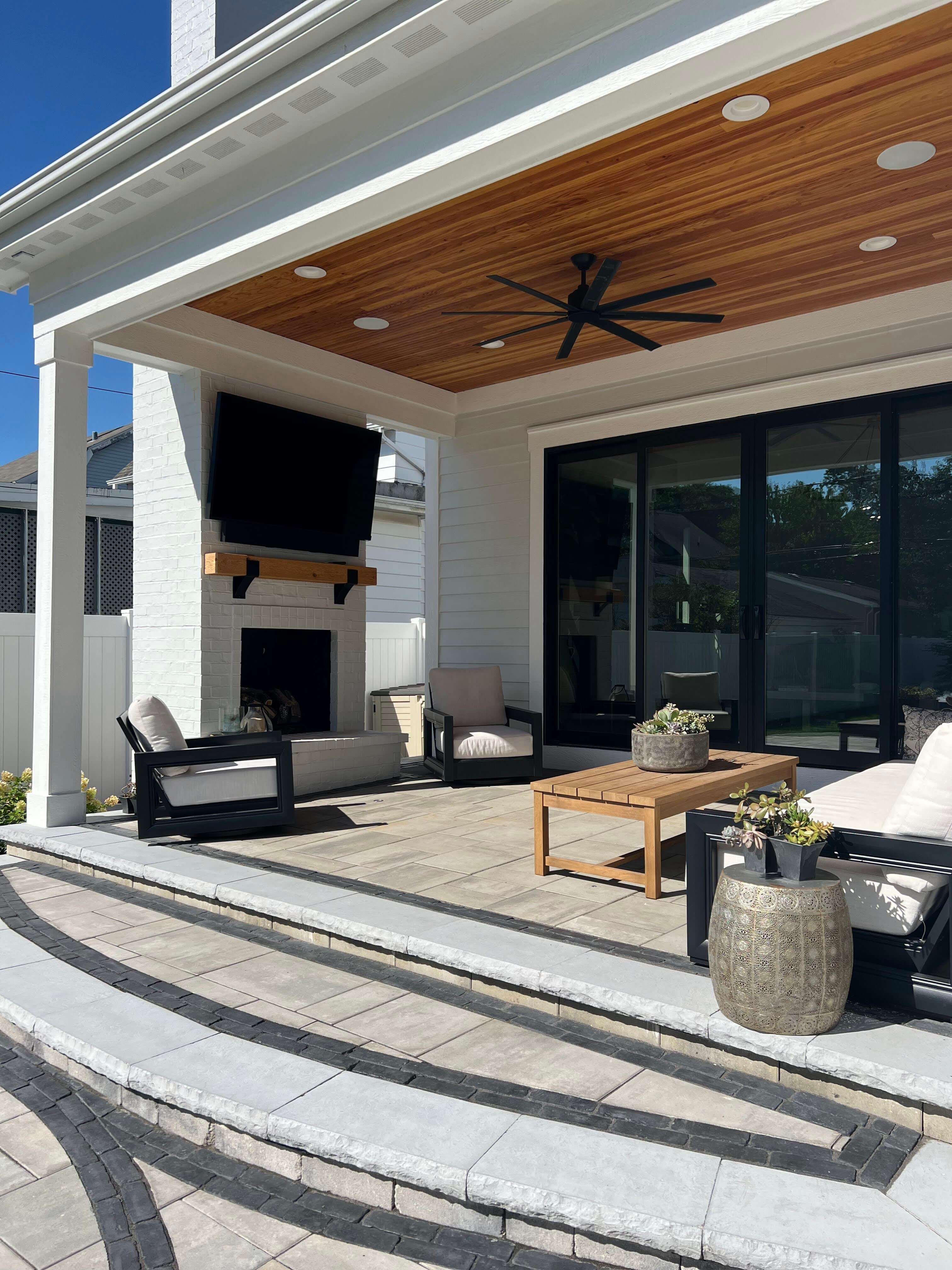 Covered patio with tile flooring, black-framed chairs with cushions, wooden coffee table, TV, and ceiling fan.
