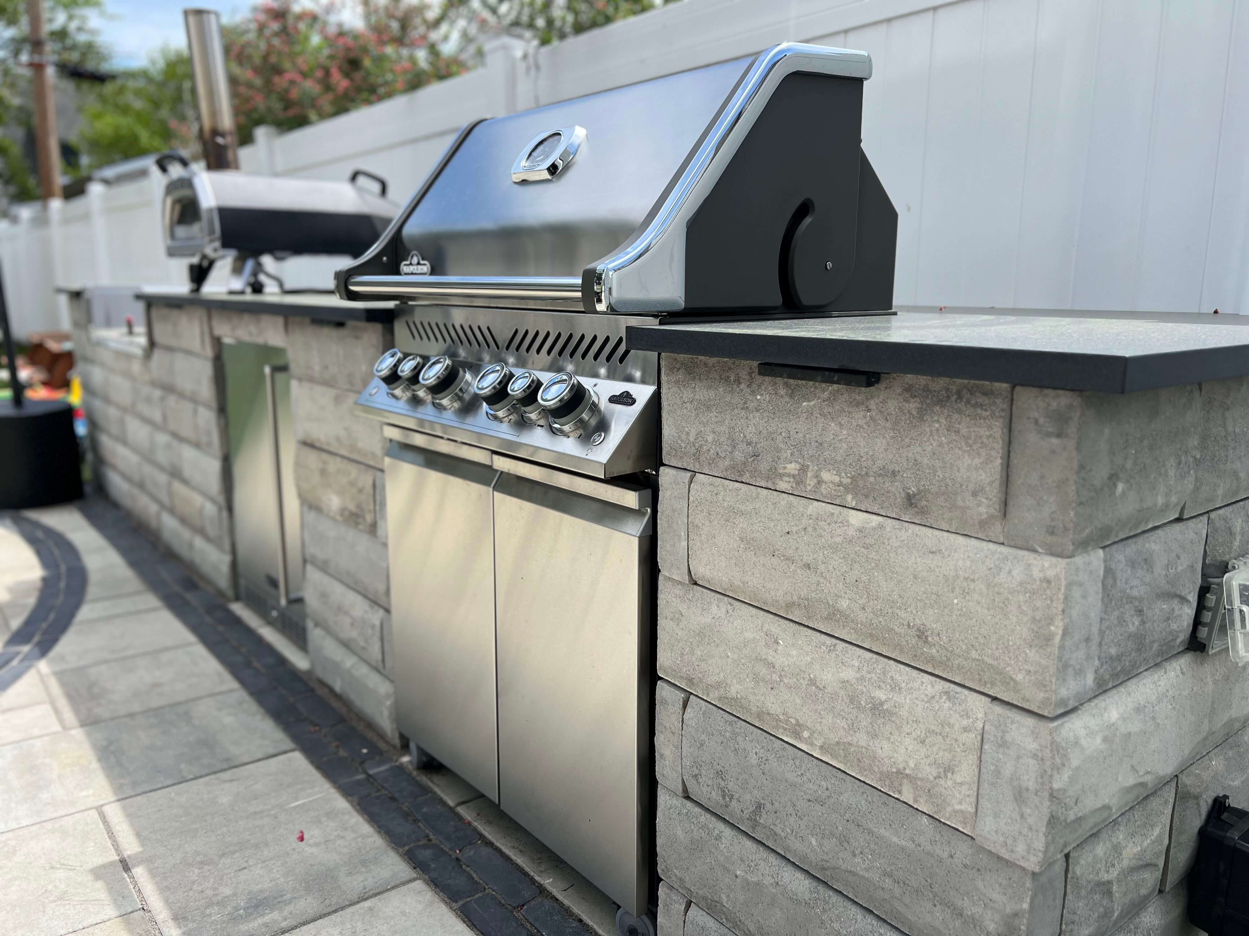 Outdoor stainless steel grill built into a gray stone countertop and patio area with white fence.