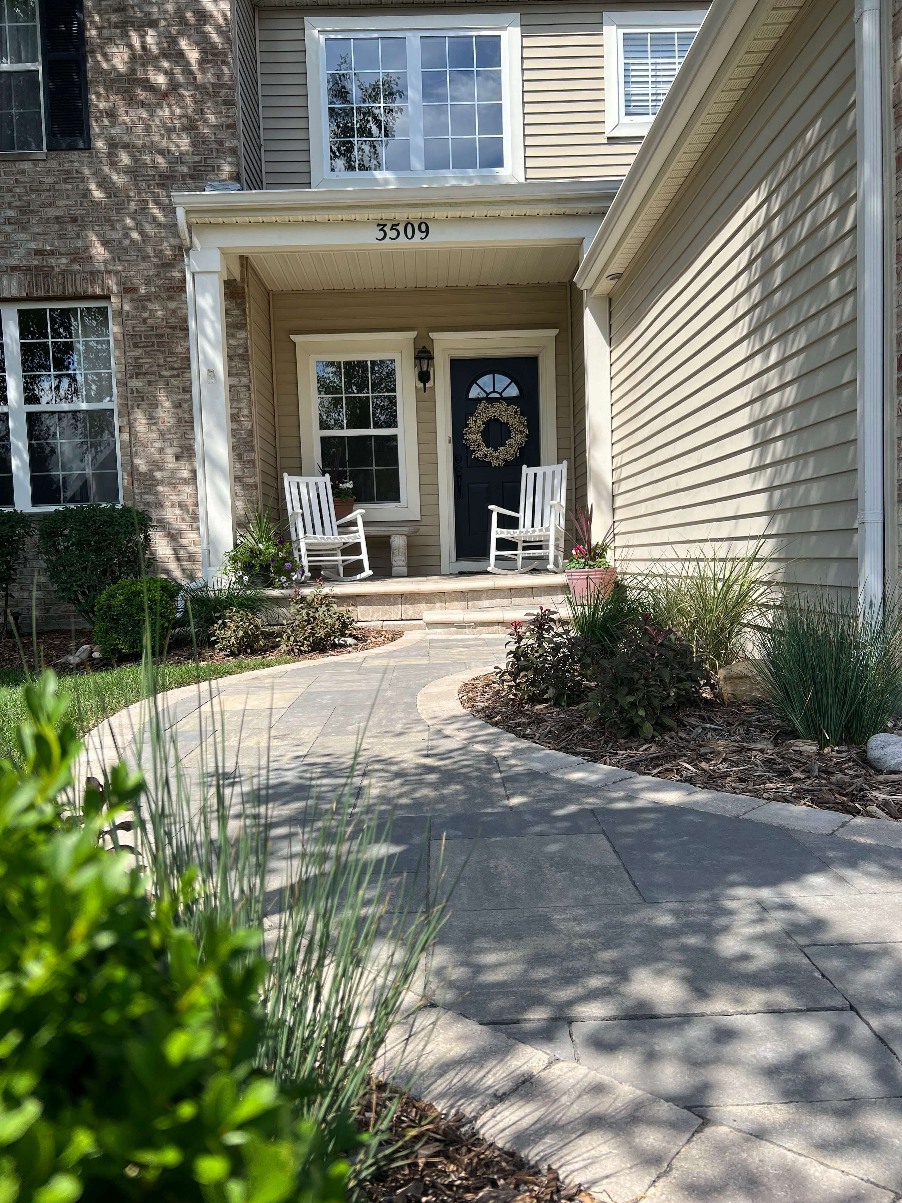 Curved stone walkway leading to a front porch with two white rocking chairs and a dark door with a wreath.
