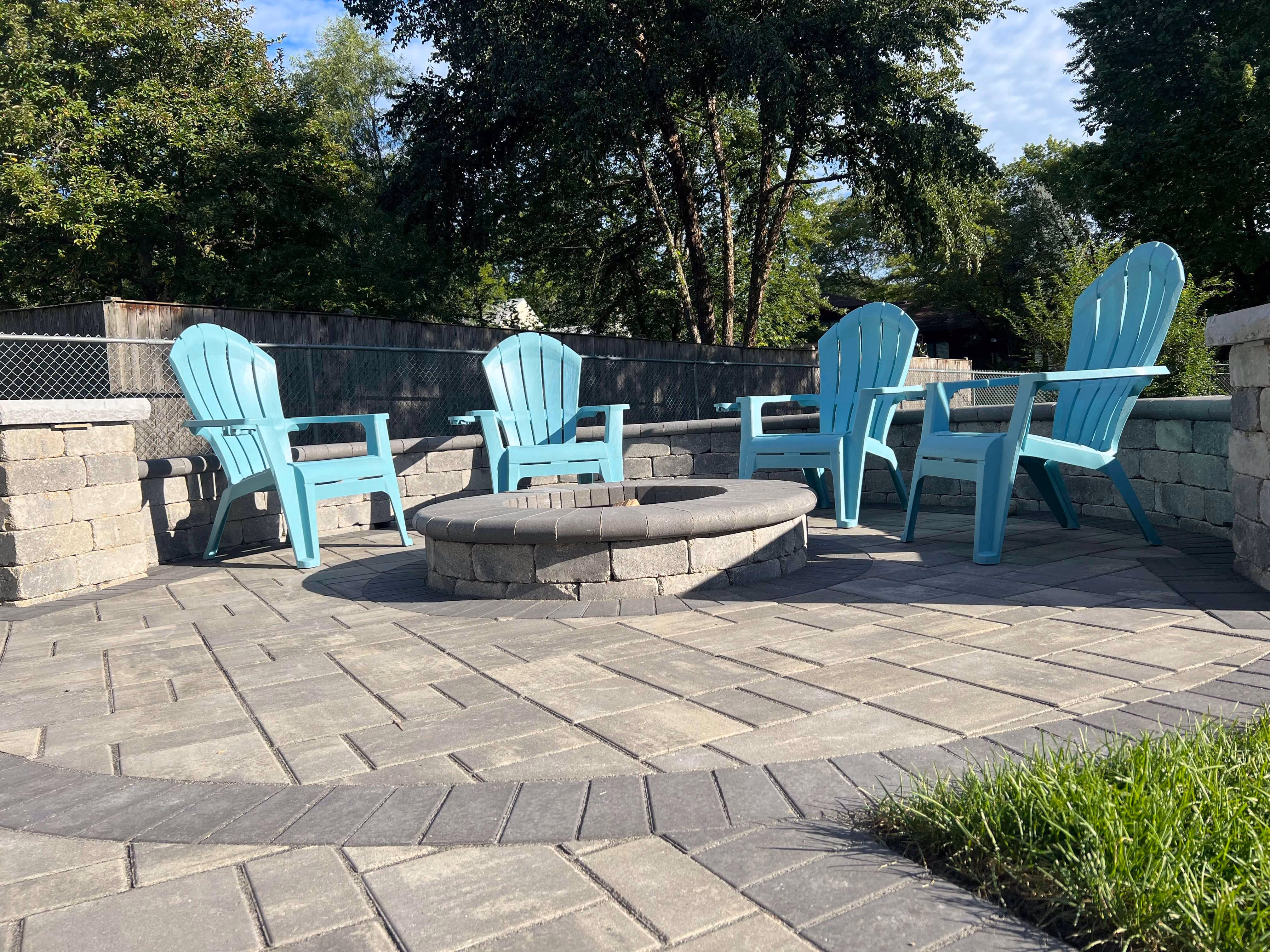 Outdoor patio with four turquoise chairs arranged around a stone fire pit on a paved surface.