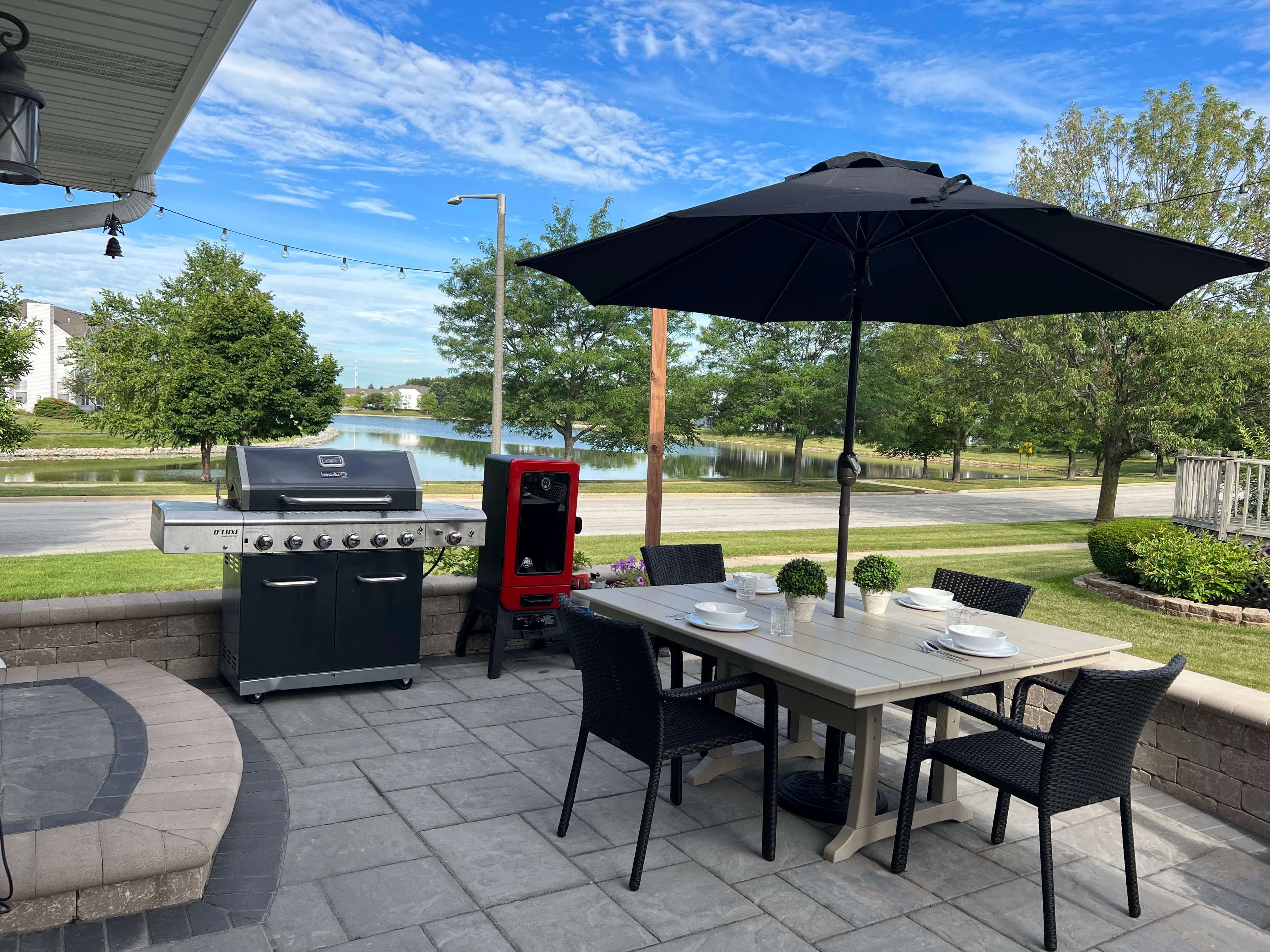 Patio dining set with black umbrella, grill, smoker, and lake with trees in background.
