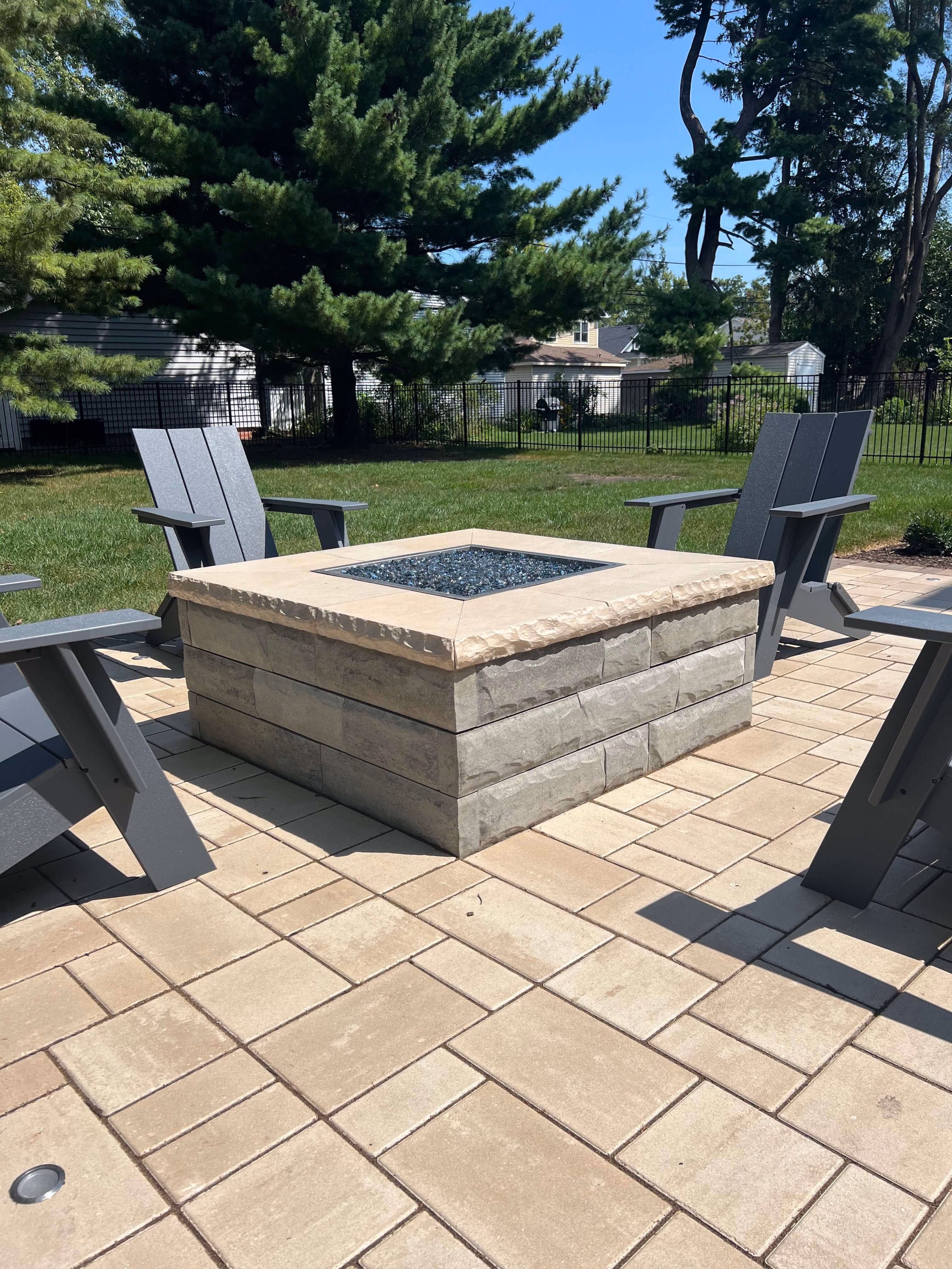 Square stone fire pit surrounded by gray Adirondack chairs on a tan brick patio in a backyard.