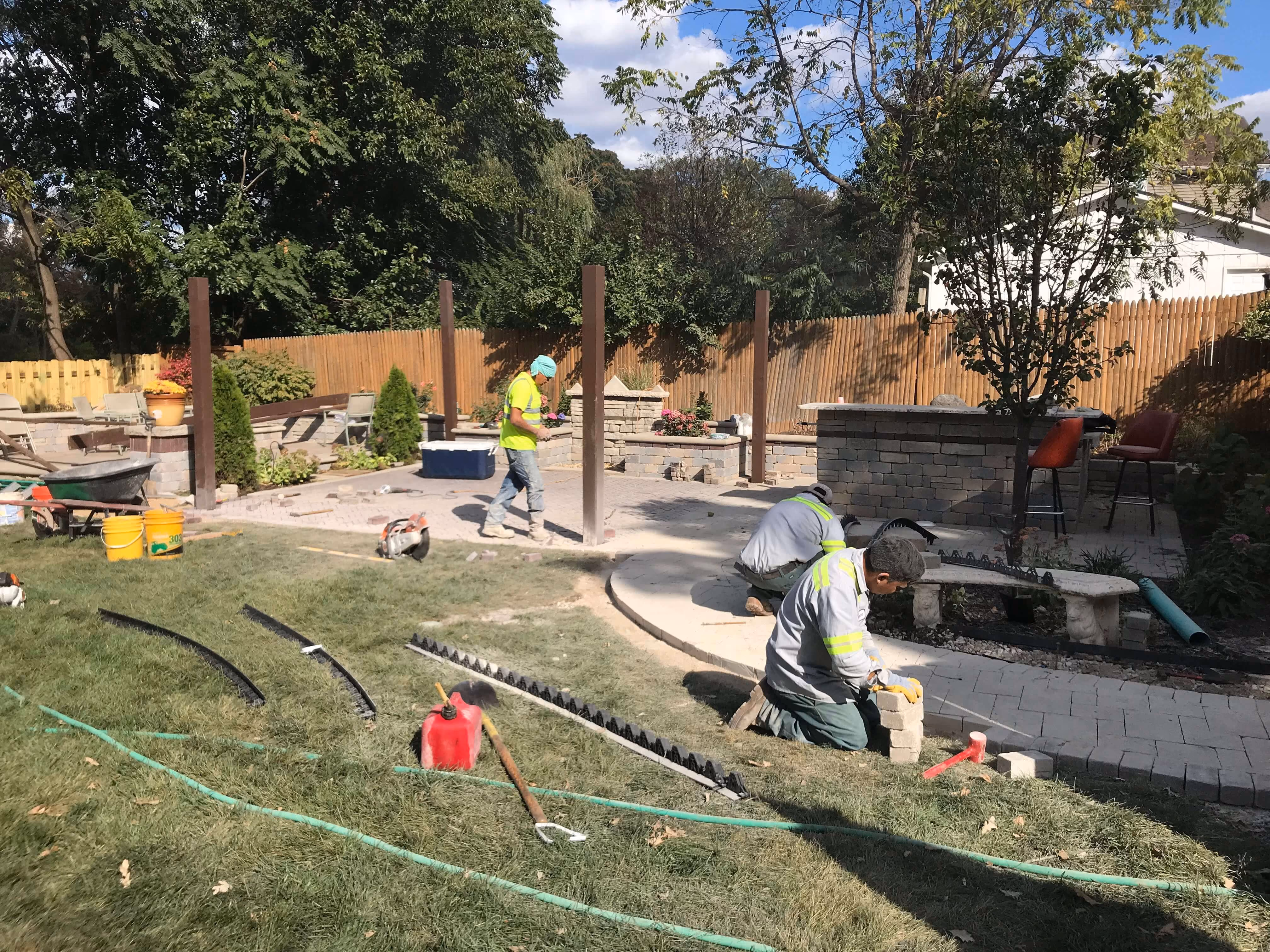Three workers installing pavers in a backyard with seating, trees, and wooden fence.