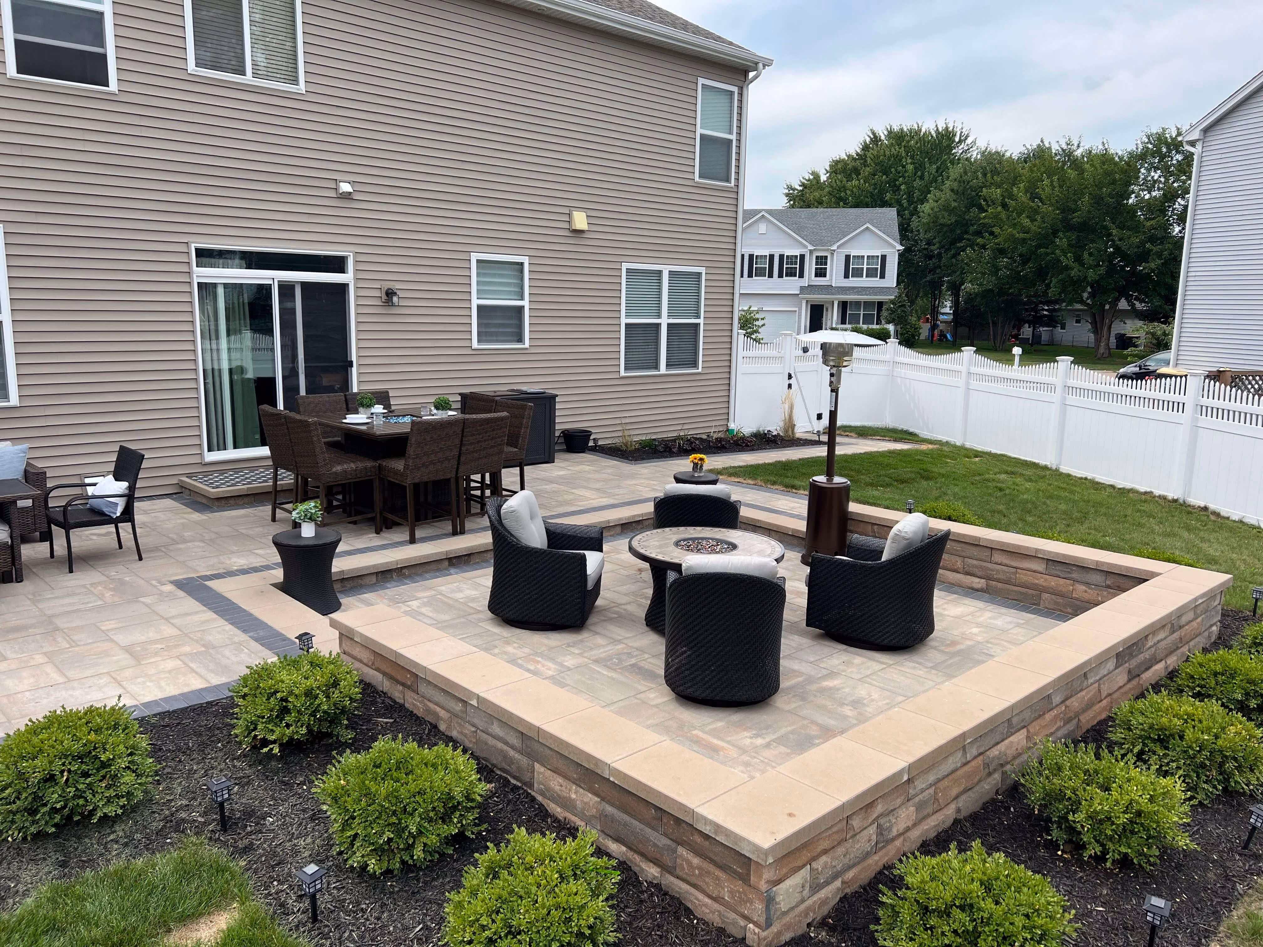 Backyard patio with sunken seating area, four black chairs, stone fire pit, and dining table near house.