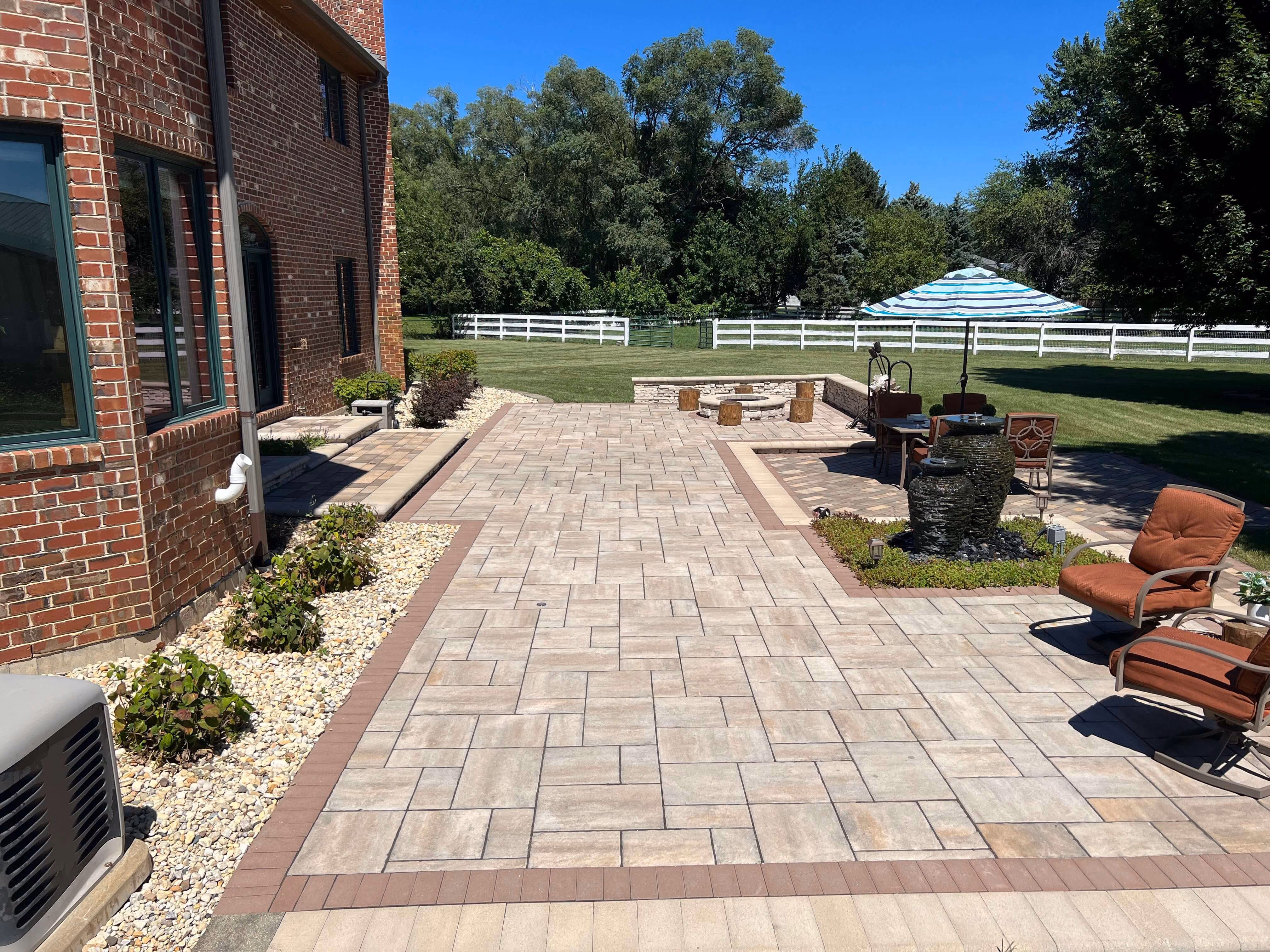 Backyard patio with beige pavers, seating area, water fountain, green lawn, and white fence under blue sky.