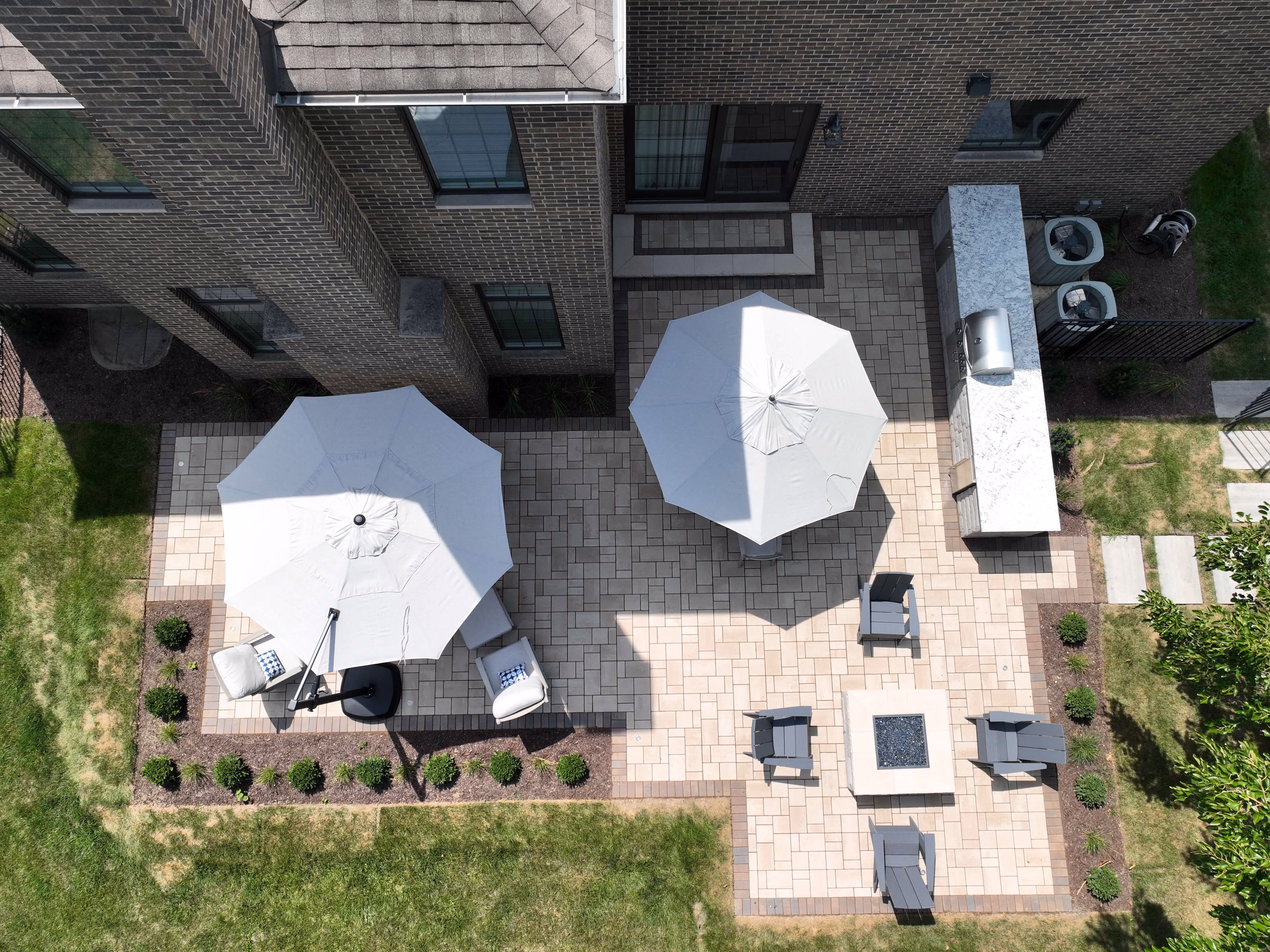 Aerial view of a brick patio with two white umbrellas, seating, a firepit, and an outdoor grill area.
