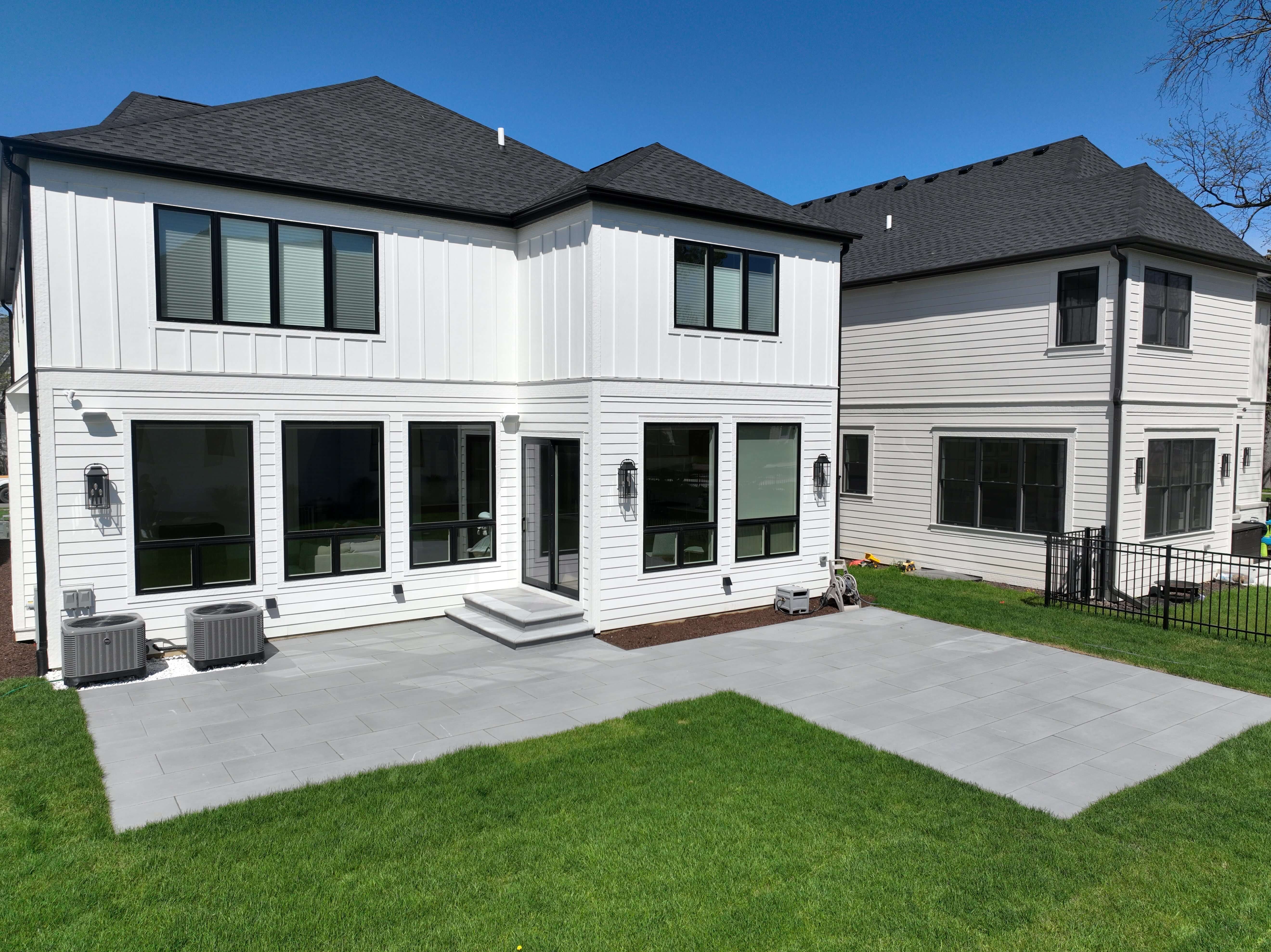 White two-story house with black windows and a spacious gray stone patio in a green backyard.