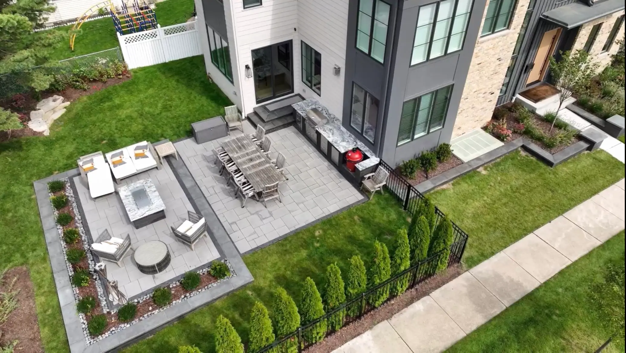Modern house backyard with patio dining set, lounge chairs, outdoor kitchen, and green lawn fenced by shrubs.