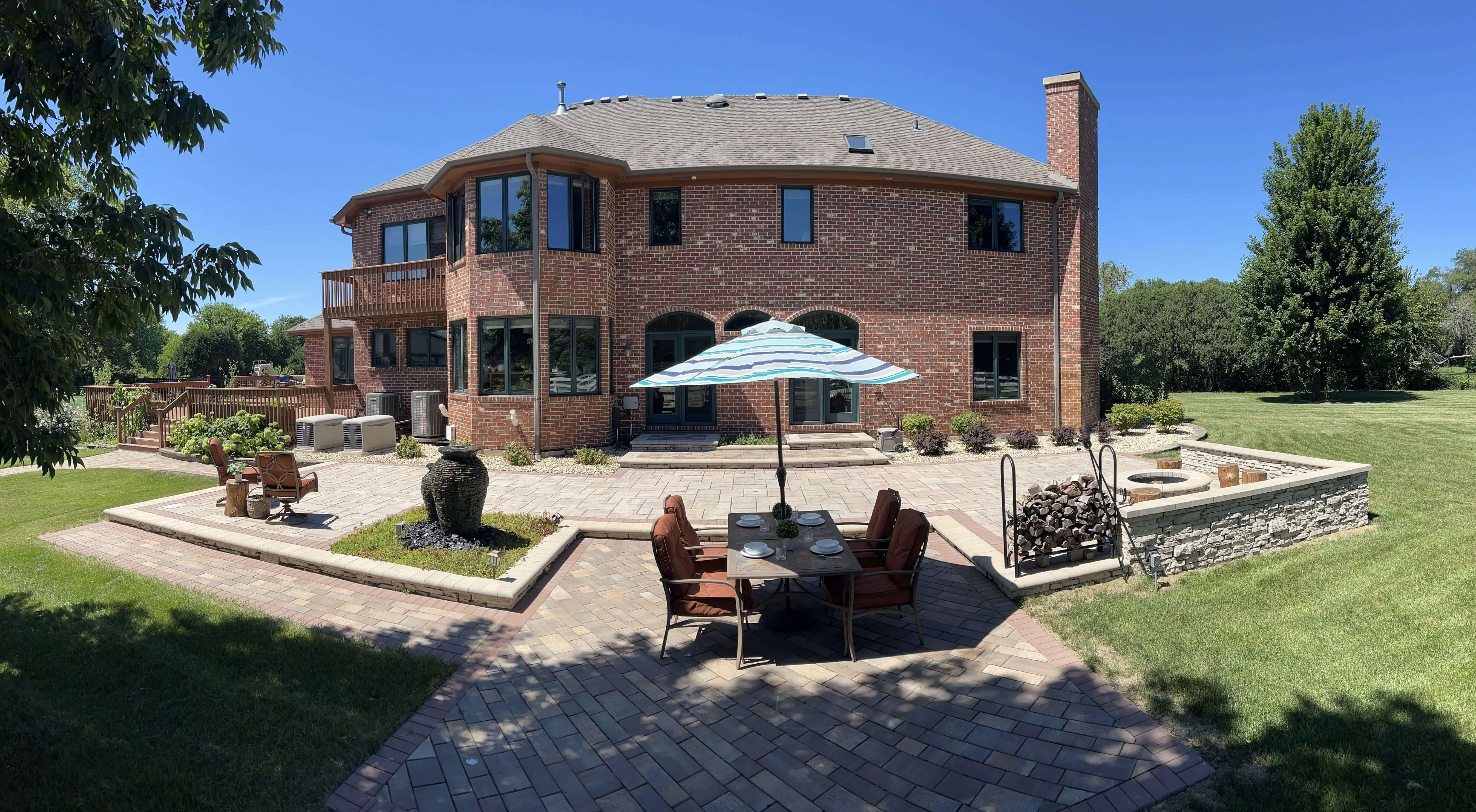 Backyard patio with table and umbrella, firewood rack, fire pit, and modern two-story red brick house.