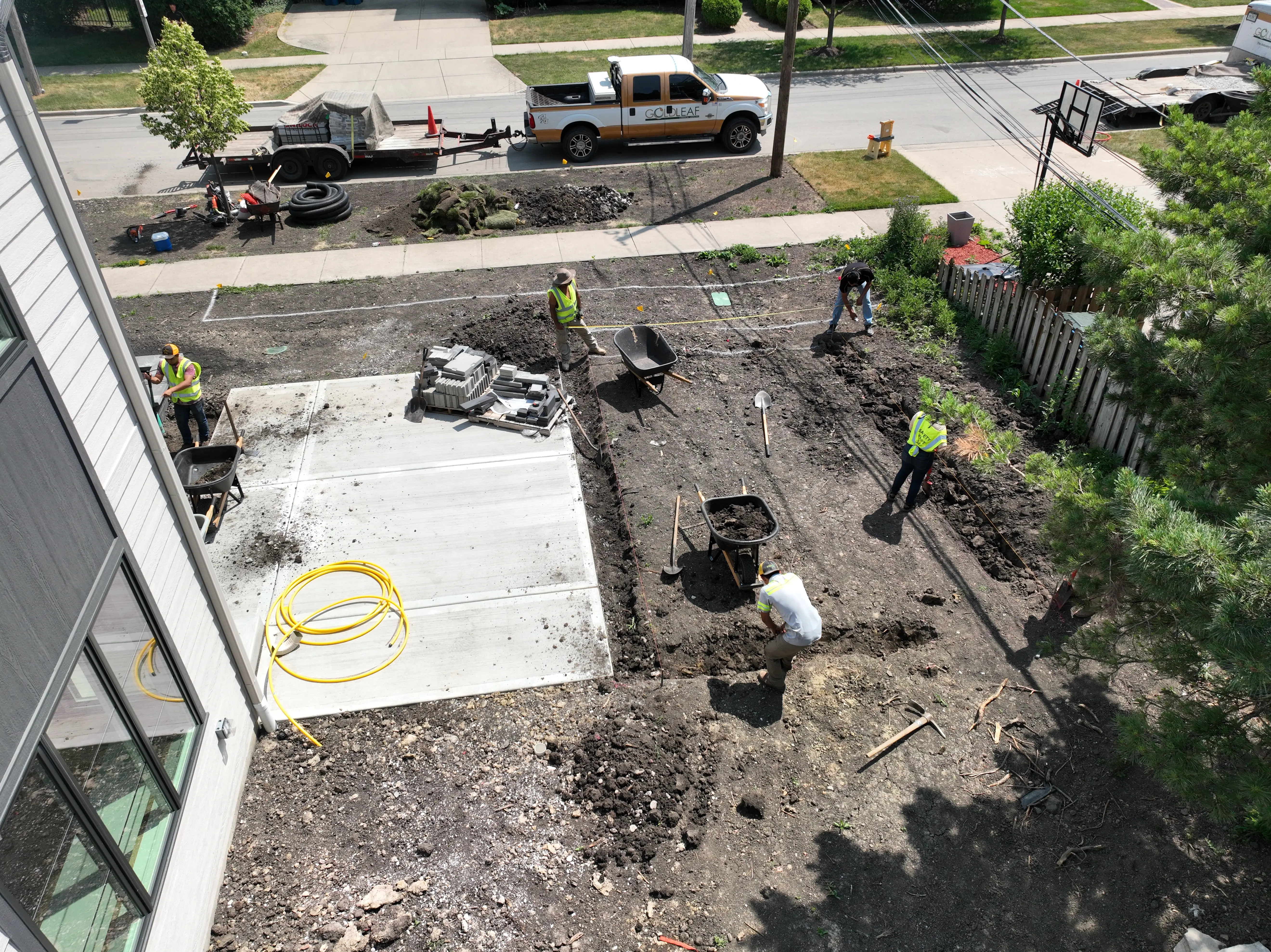 Workers in safety vests preparing a front yard with shovels, wheelbarrows, and construction materials.