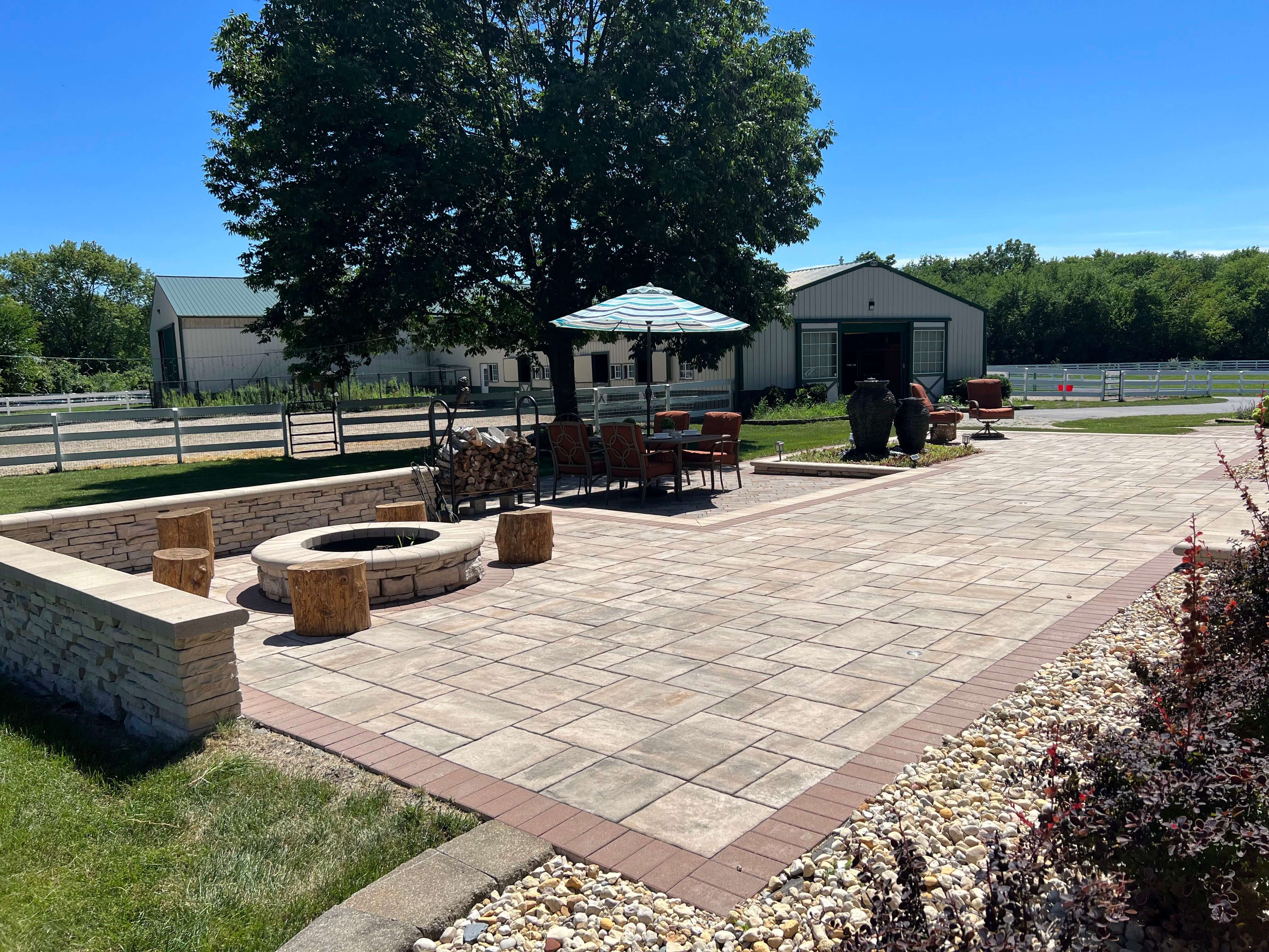 Spacious backyard patio with stone fire pit, wooden stools, seating area, umbrella, and barn in background.