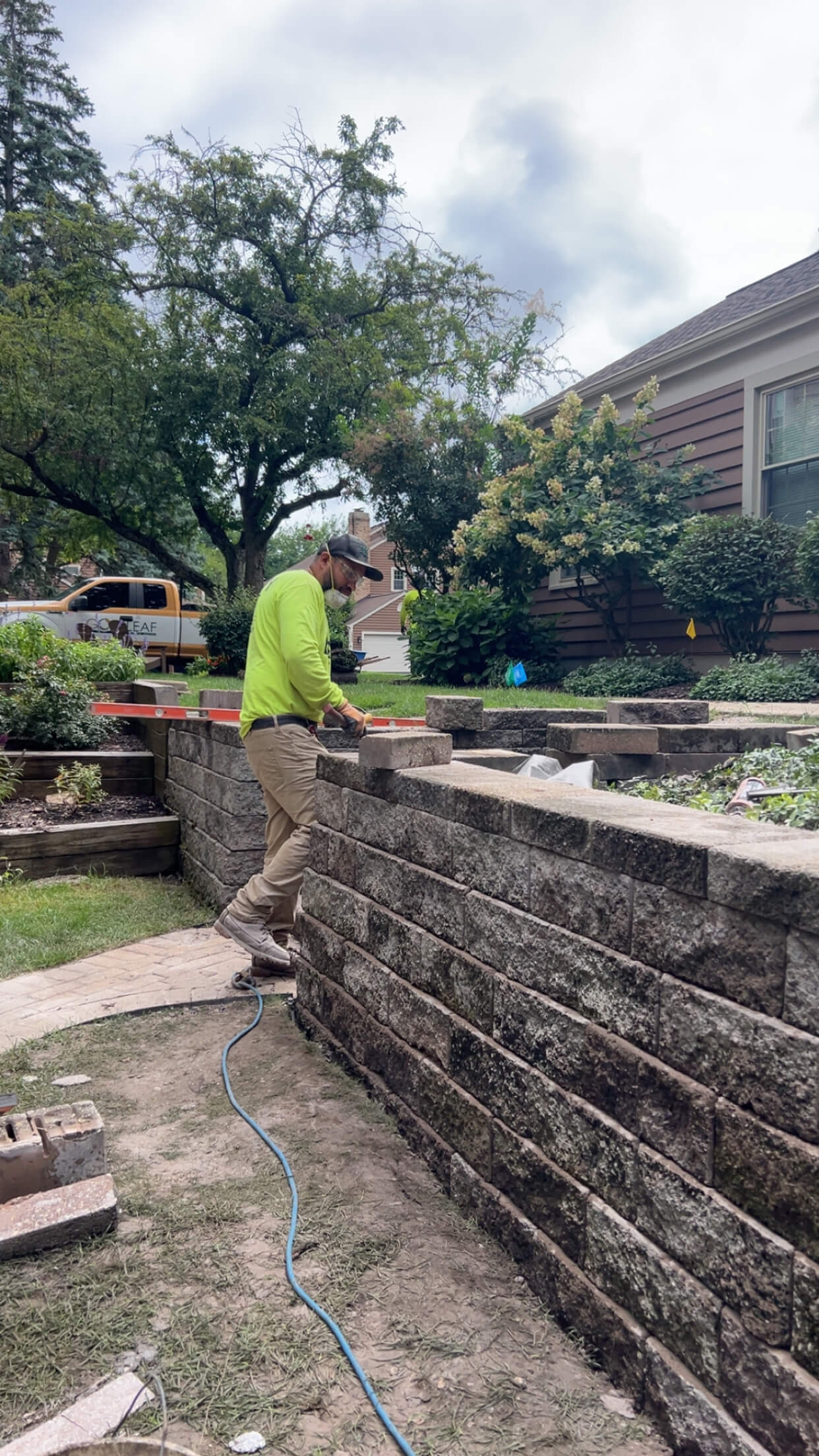 Worker in a yellow shirt building a stone retaining wall in a residential backyard with trees and shrubs.
