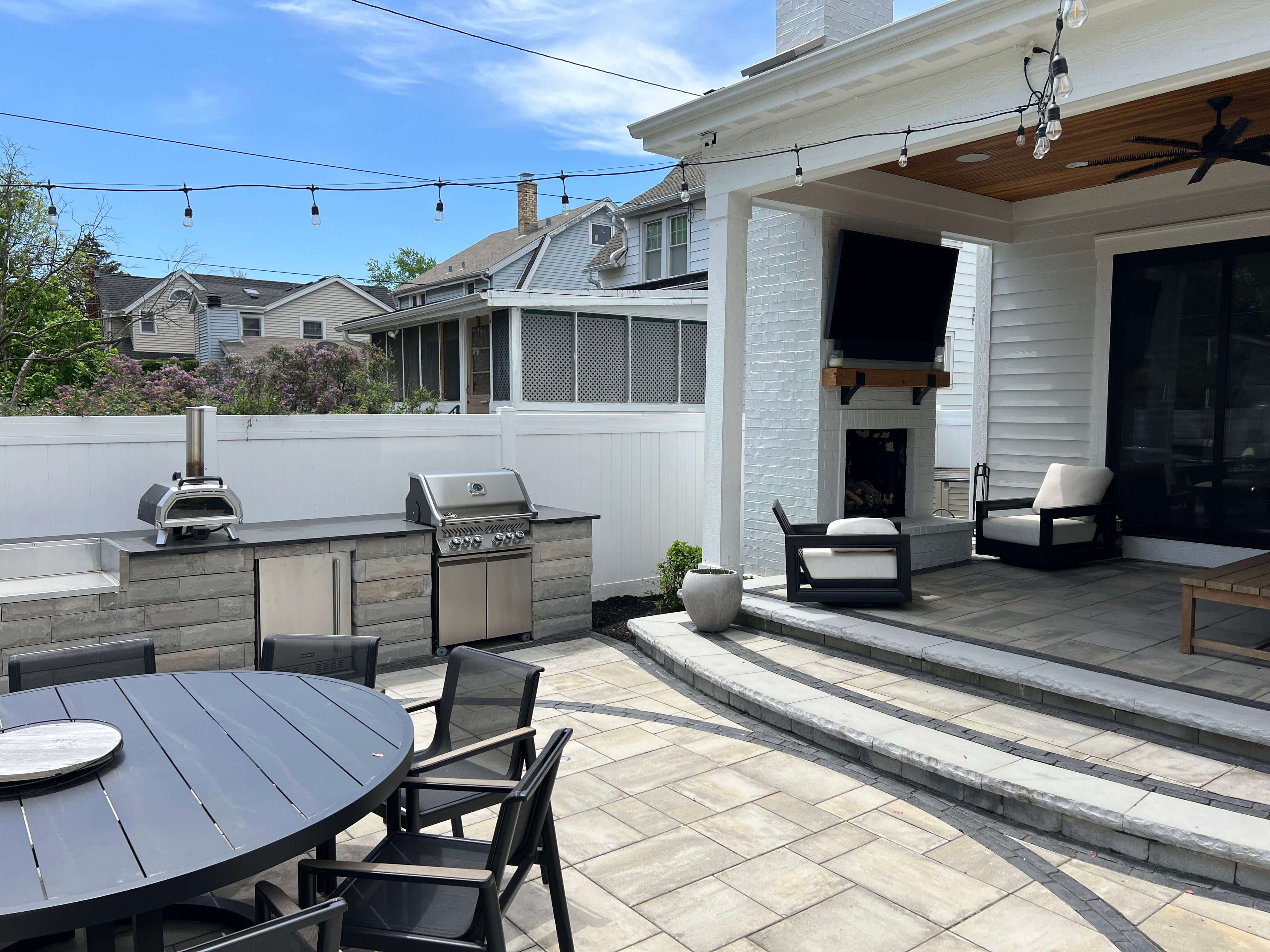 Backyard patio with outdoor kitchen, black round table with chairs, white fence, and seating area with TV.