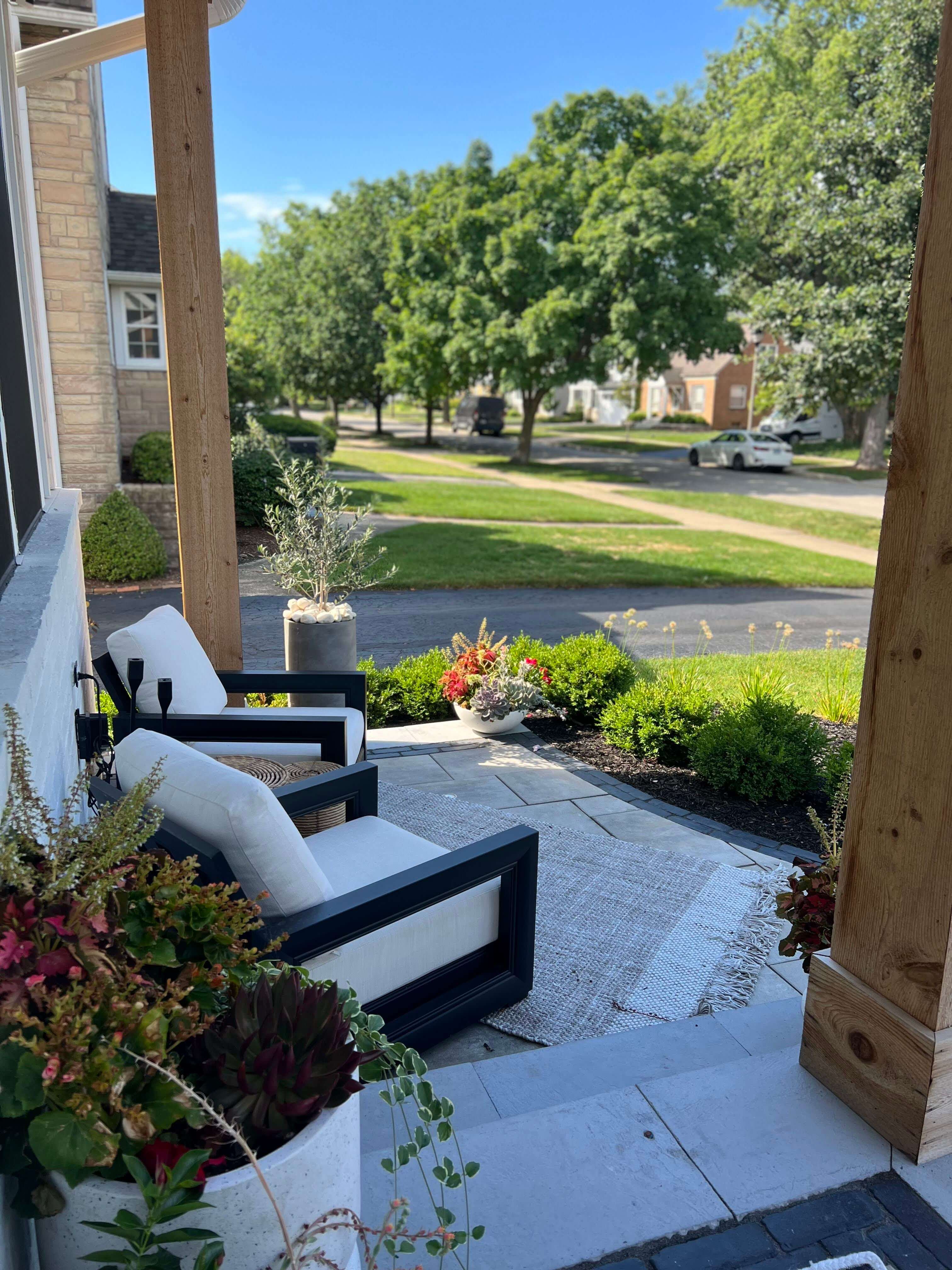 Front porch with cushioned black-frame chairs, outdoor rug, potted plants, and a suburban street view.