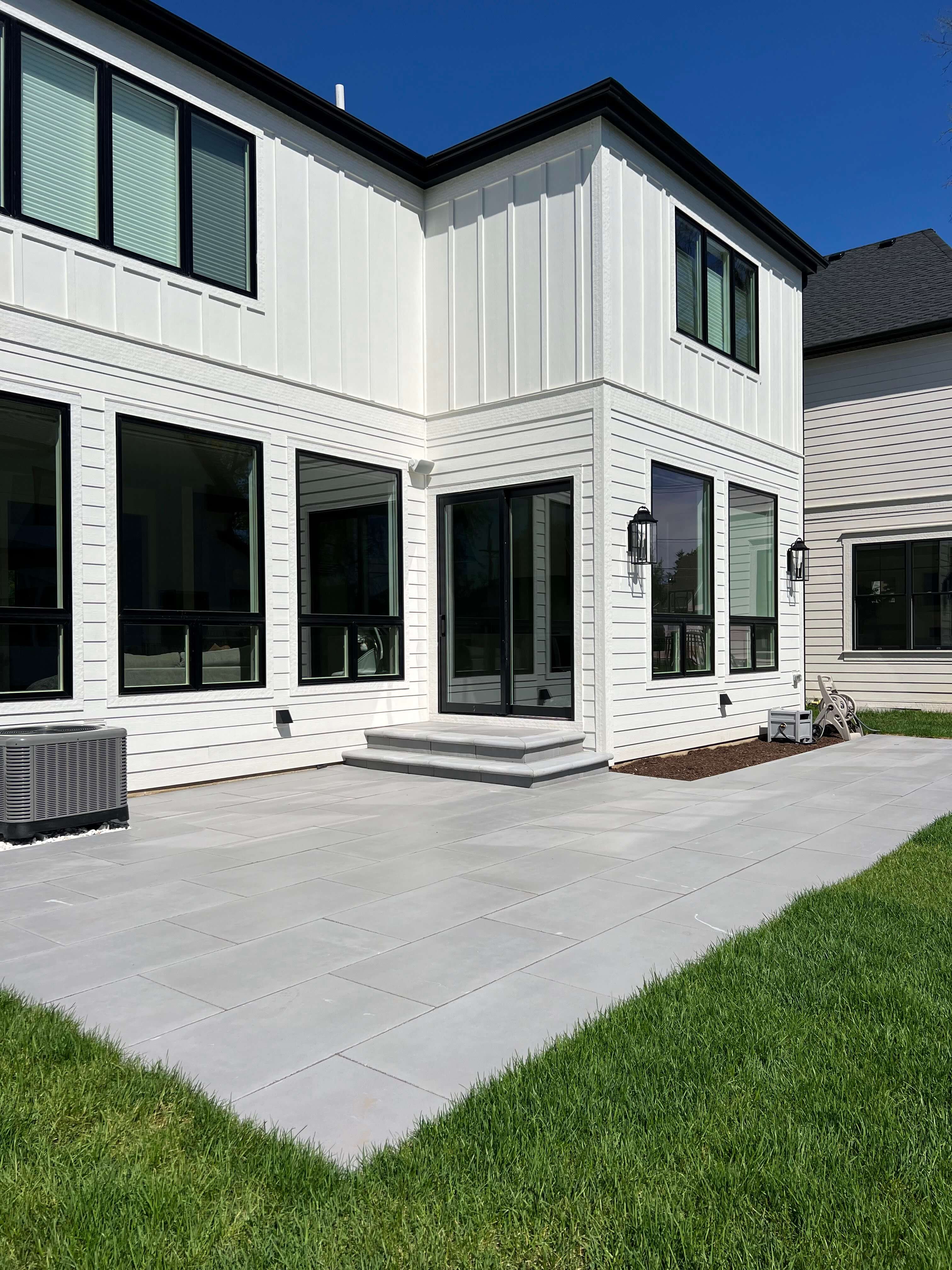Modern white house with black-framed windows and glass doors opening to a gray tiled patio and green lawn.