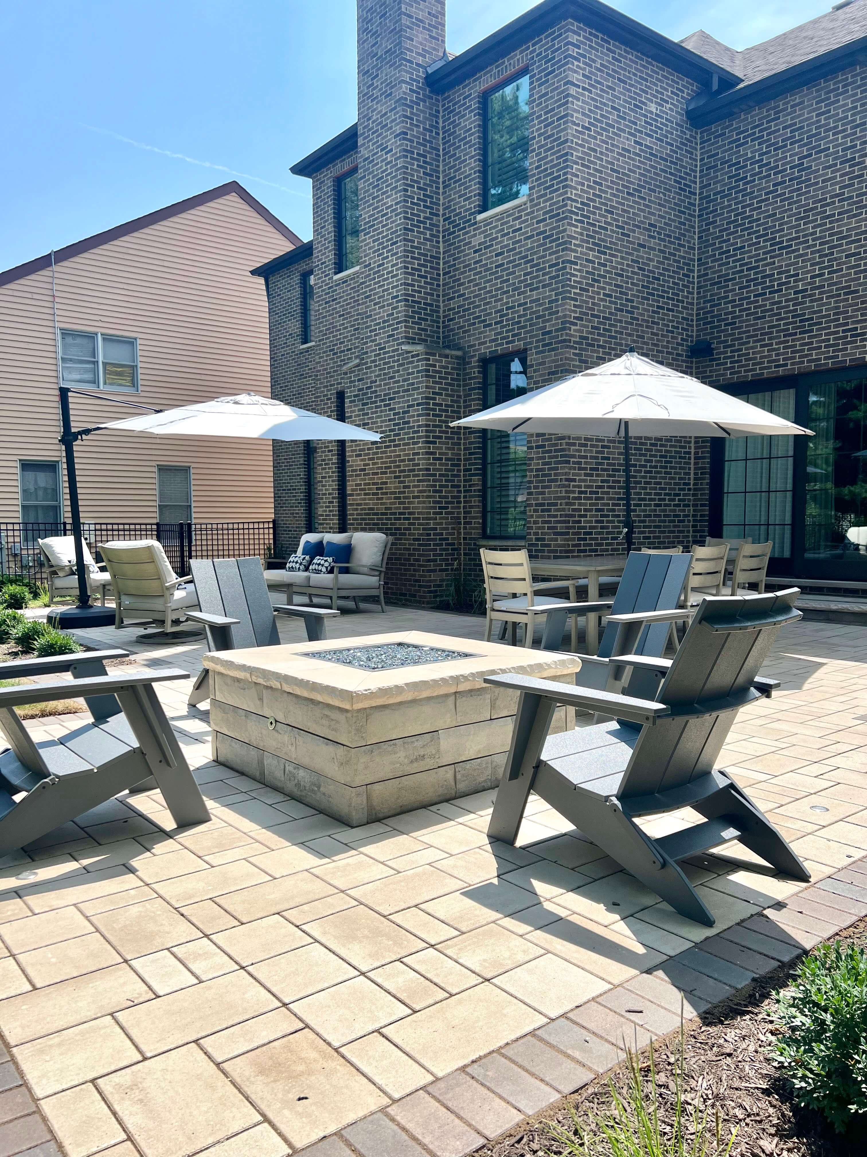 Backyard patio with stone fire pit, gray chairs, tables, white umbrellas, and brick house background.
