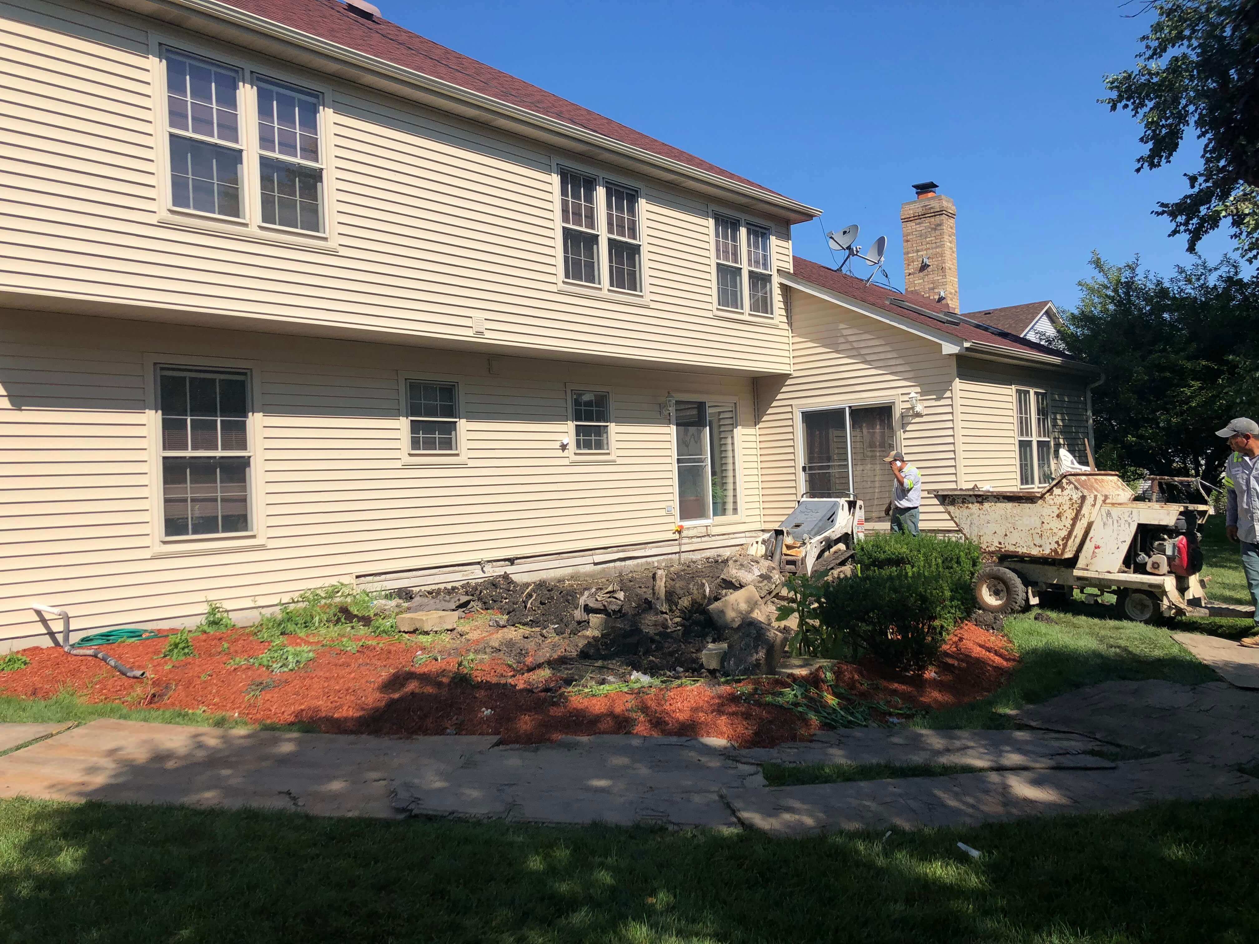 Two workers with construction equipment removing soil next to a beige house under clear blue sky.