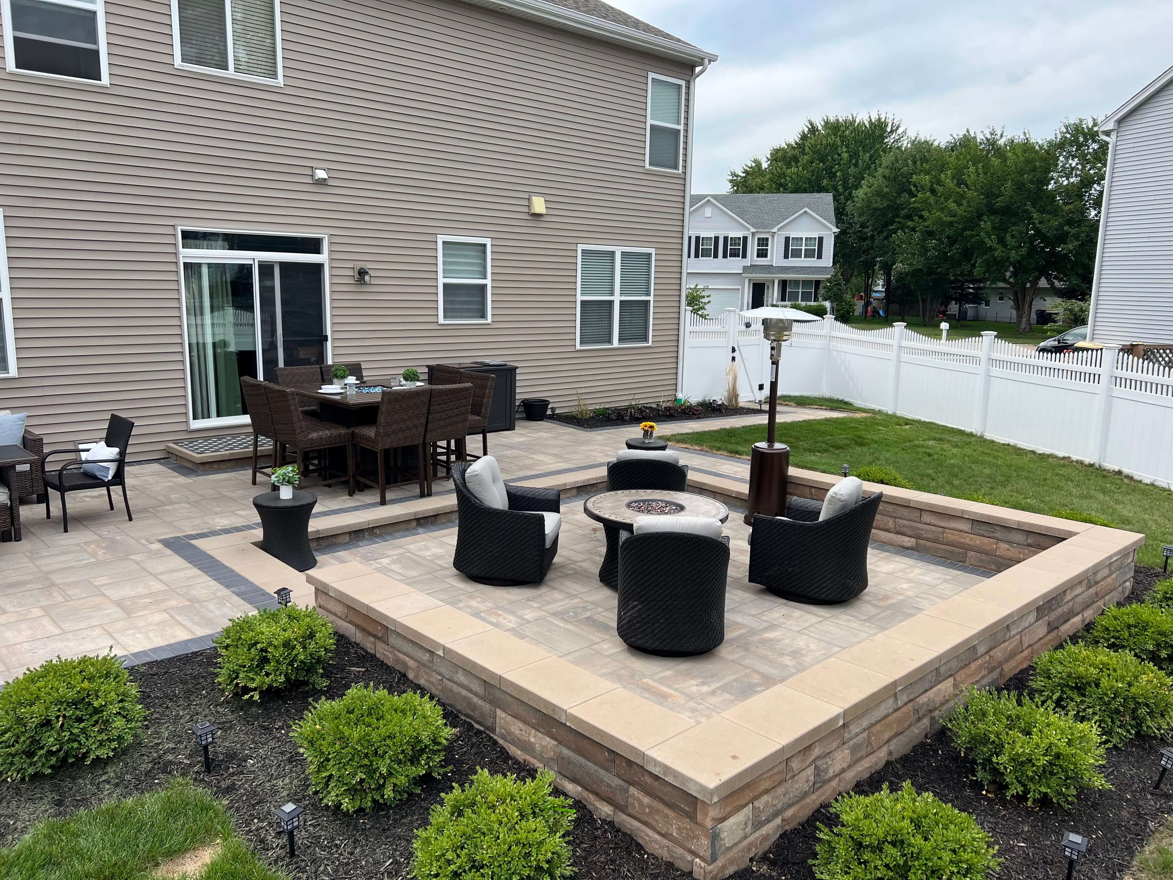 Backyard patio with sunken seating area, round fire pit table, chairs, and dining table near house.