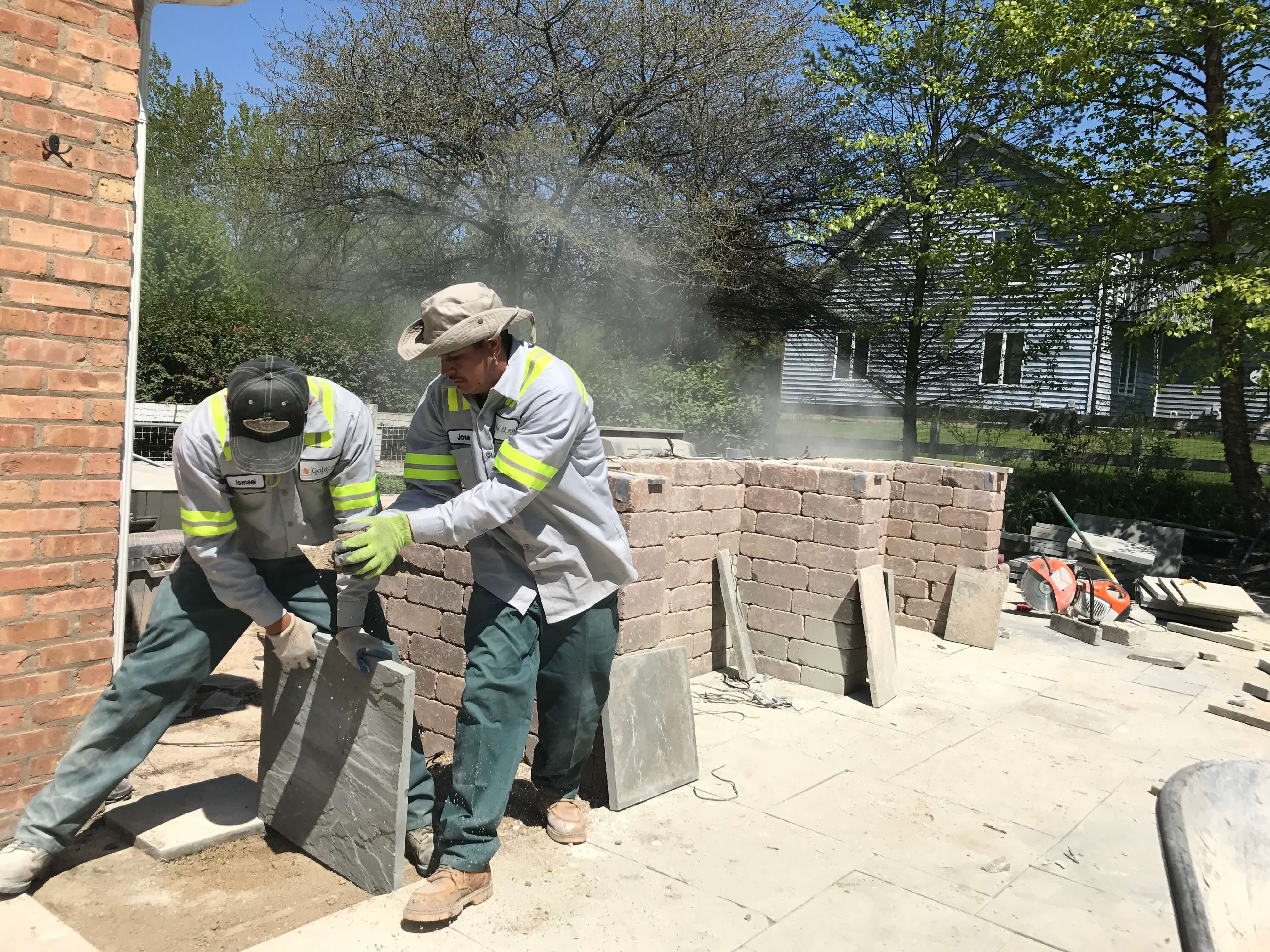 Two workers in uniforms setting large stone tiles on a patio under clear sky with trees and house in background.