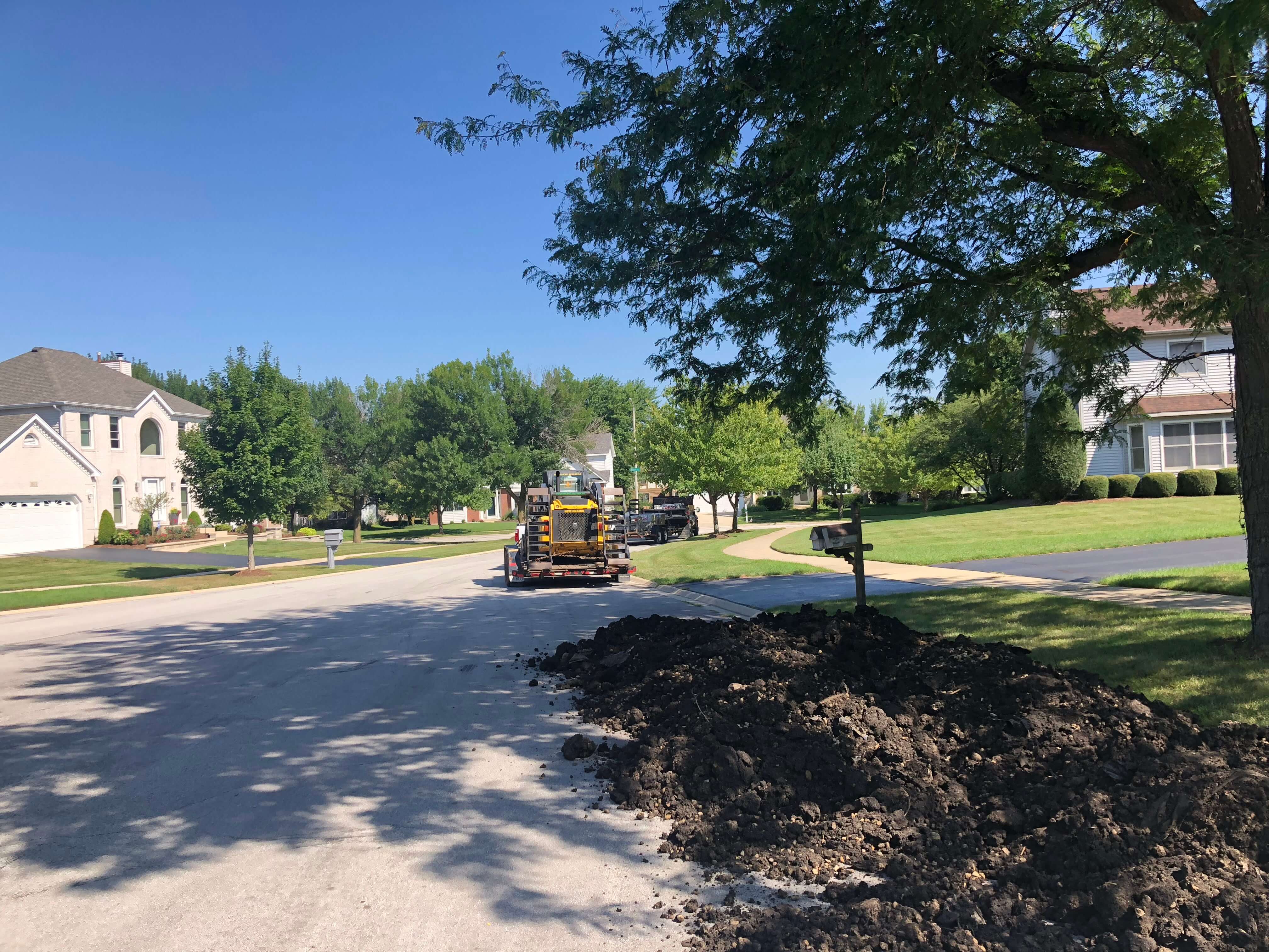 Pile of dark soil on a suburban street next to a mailbox with houses and trees under clear blue sky.