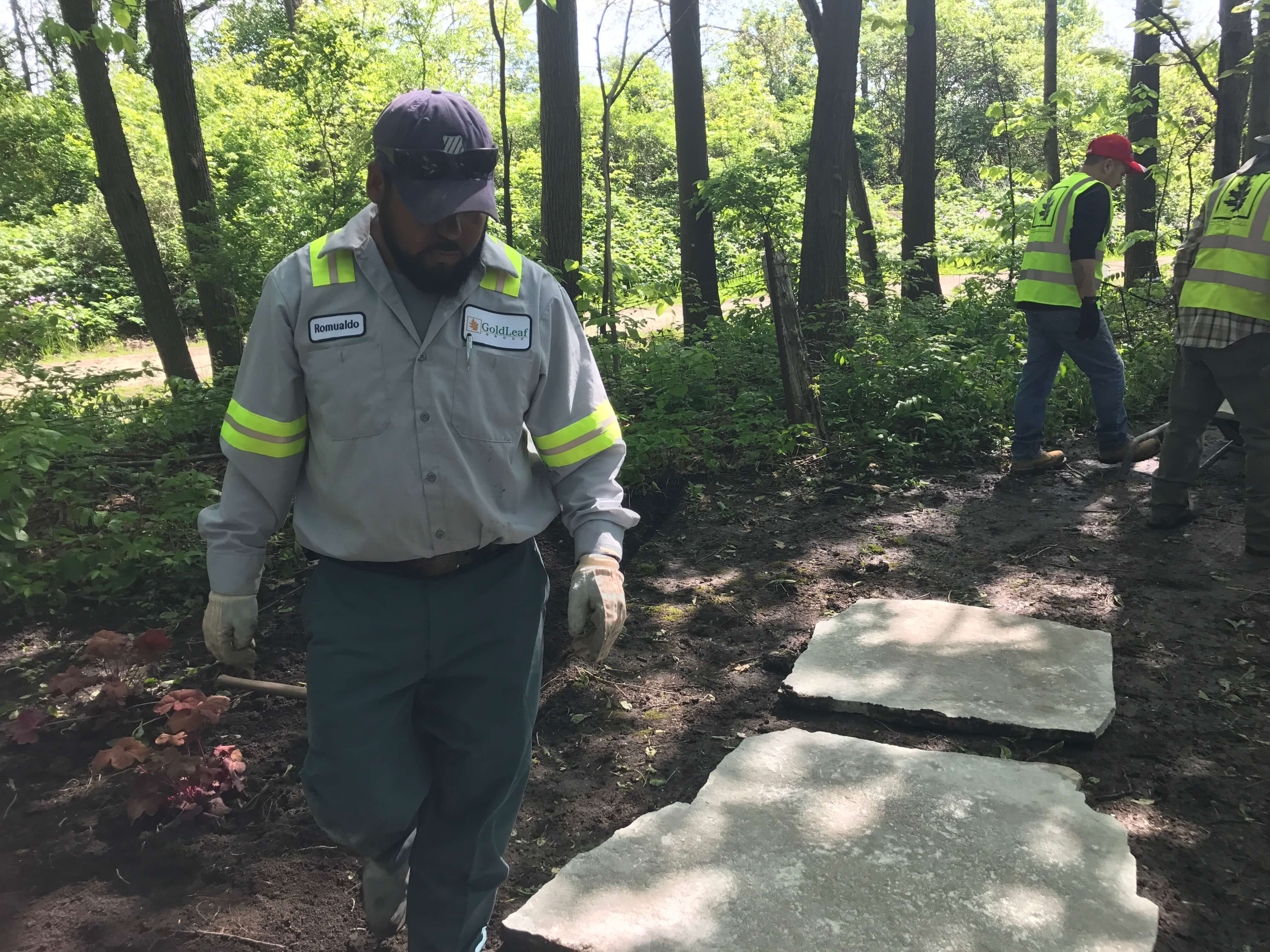Worker in uniform with GoldLeaf logo walking near stone slabs in wooded outdoor landscape area.
