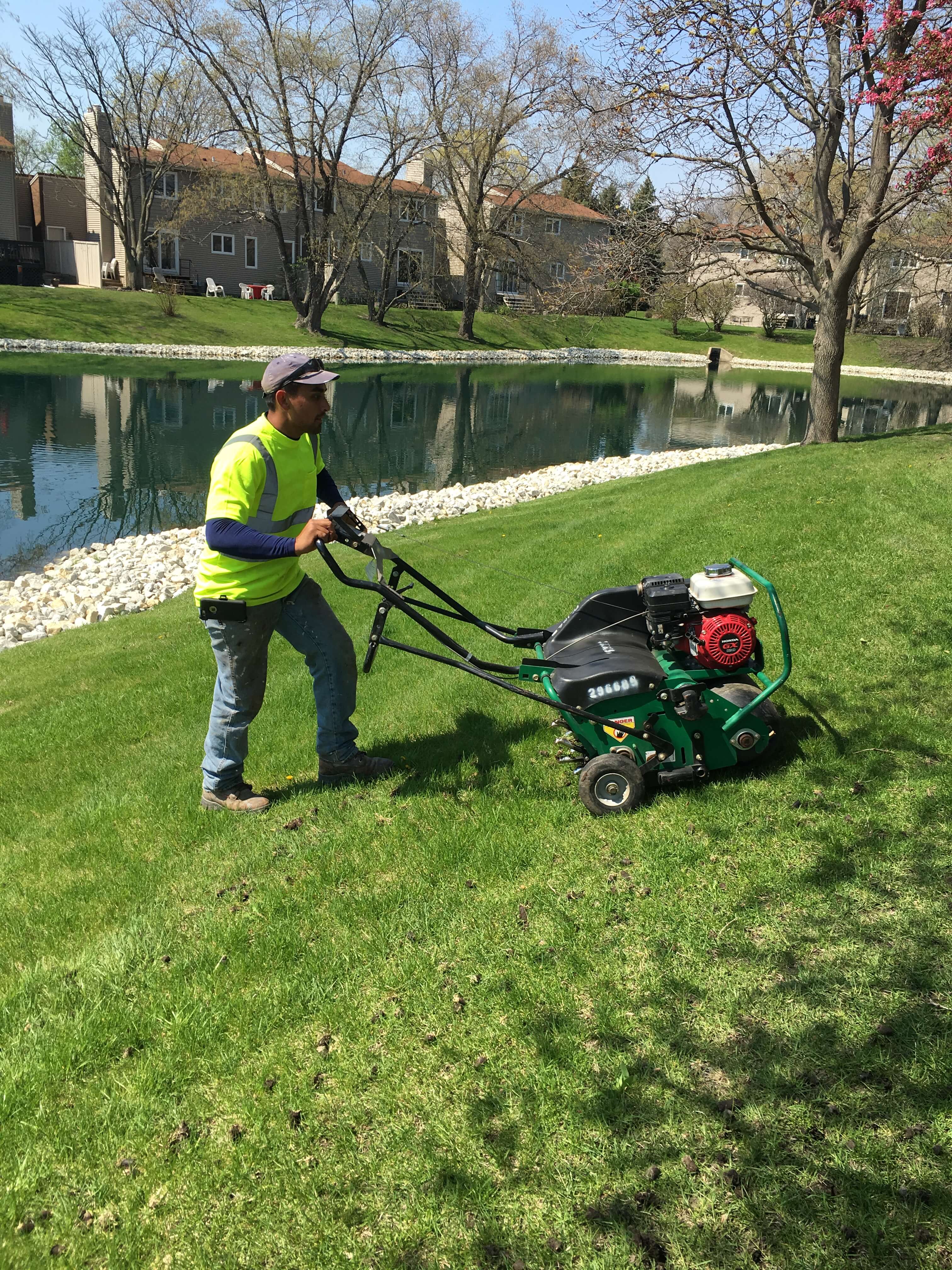 Worker in neon shirt using a green aerator machine on a green lawn near a water pond with houses in background.