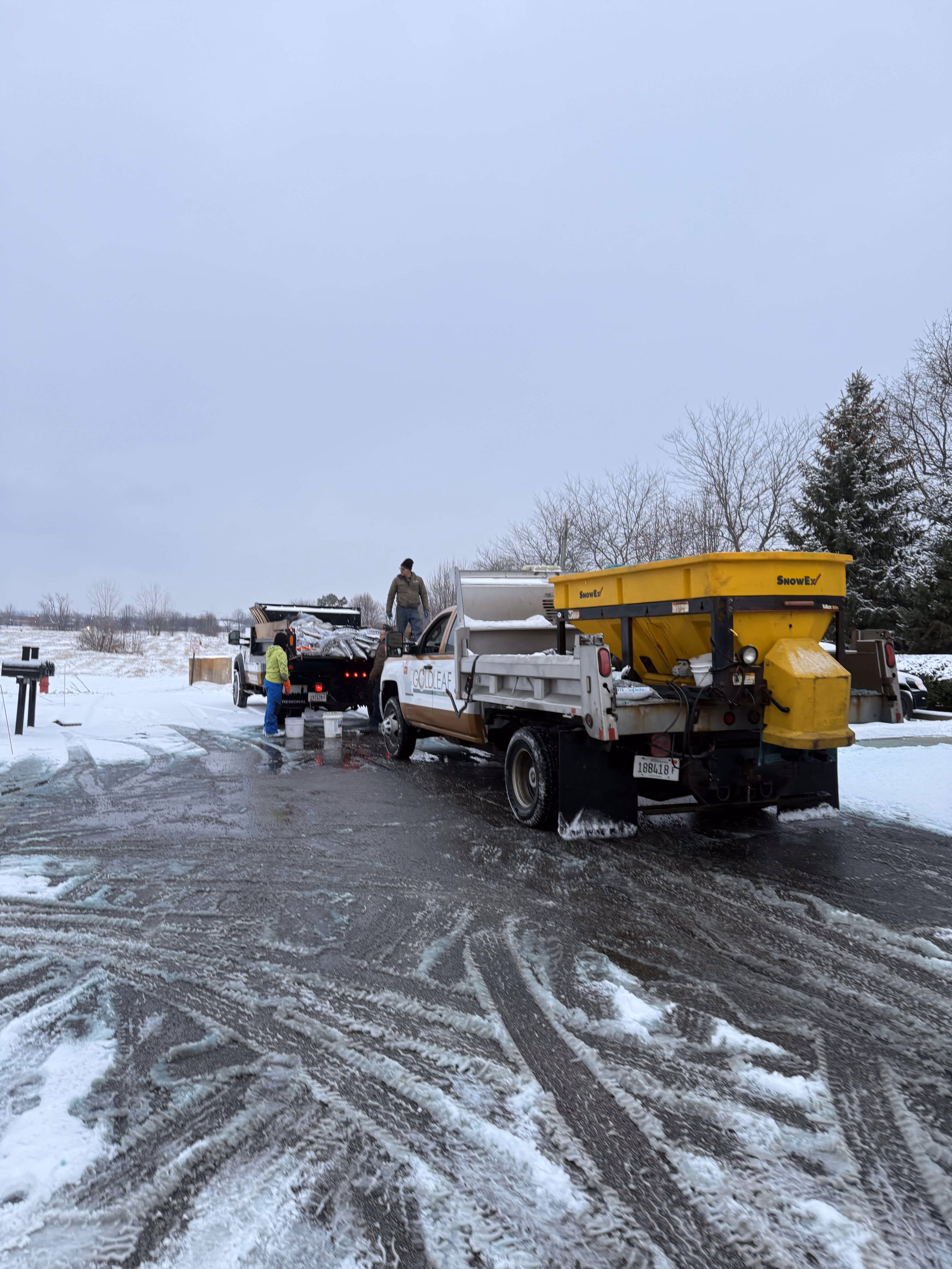Two trucks with snow removal equipment and workers handling supplies on a snowy, icy road.