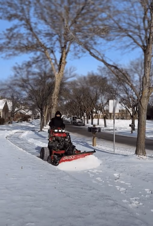 Person riding a red snowplow clearing a snowy sidewalk on a residential street with bare trees.