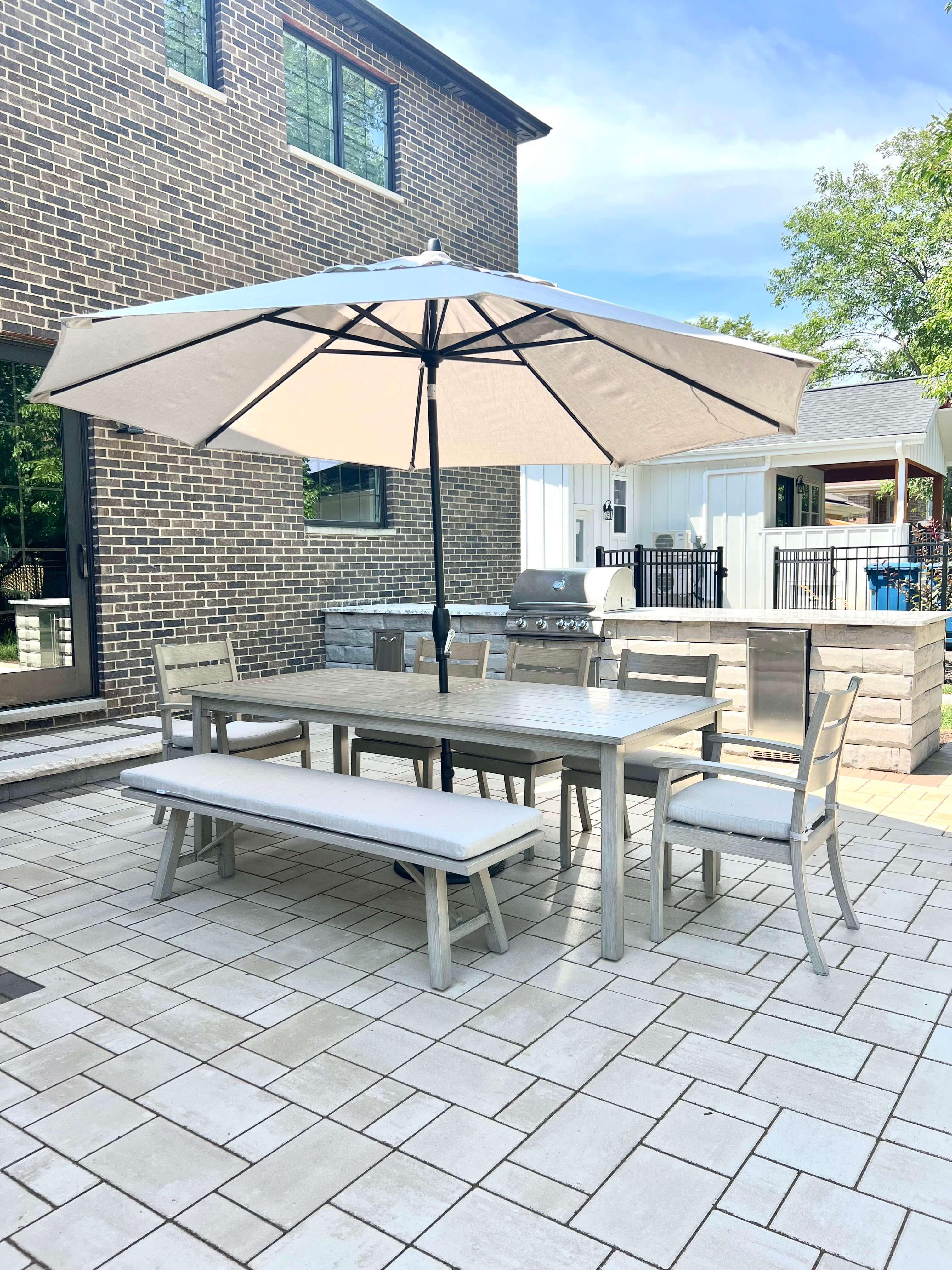 Outdoor patio with beige umbrella shading dining table, chairs, bench, and stainless steel grill.