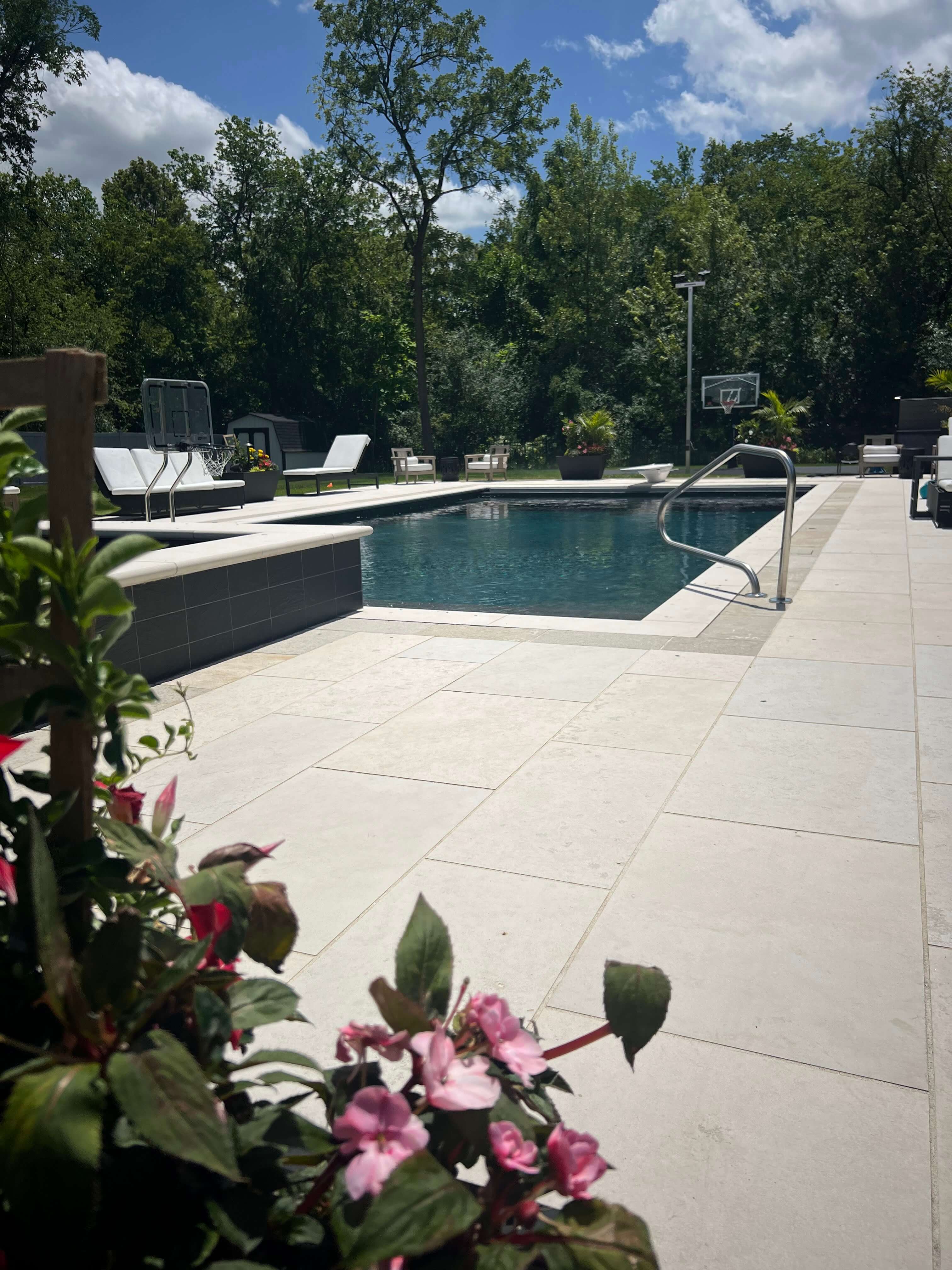 Backyard pool with white tile deck, lounge chairs, basketball hoop, and trees in the background.