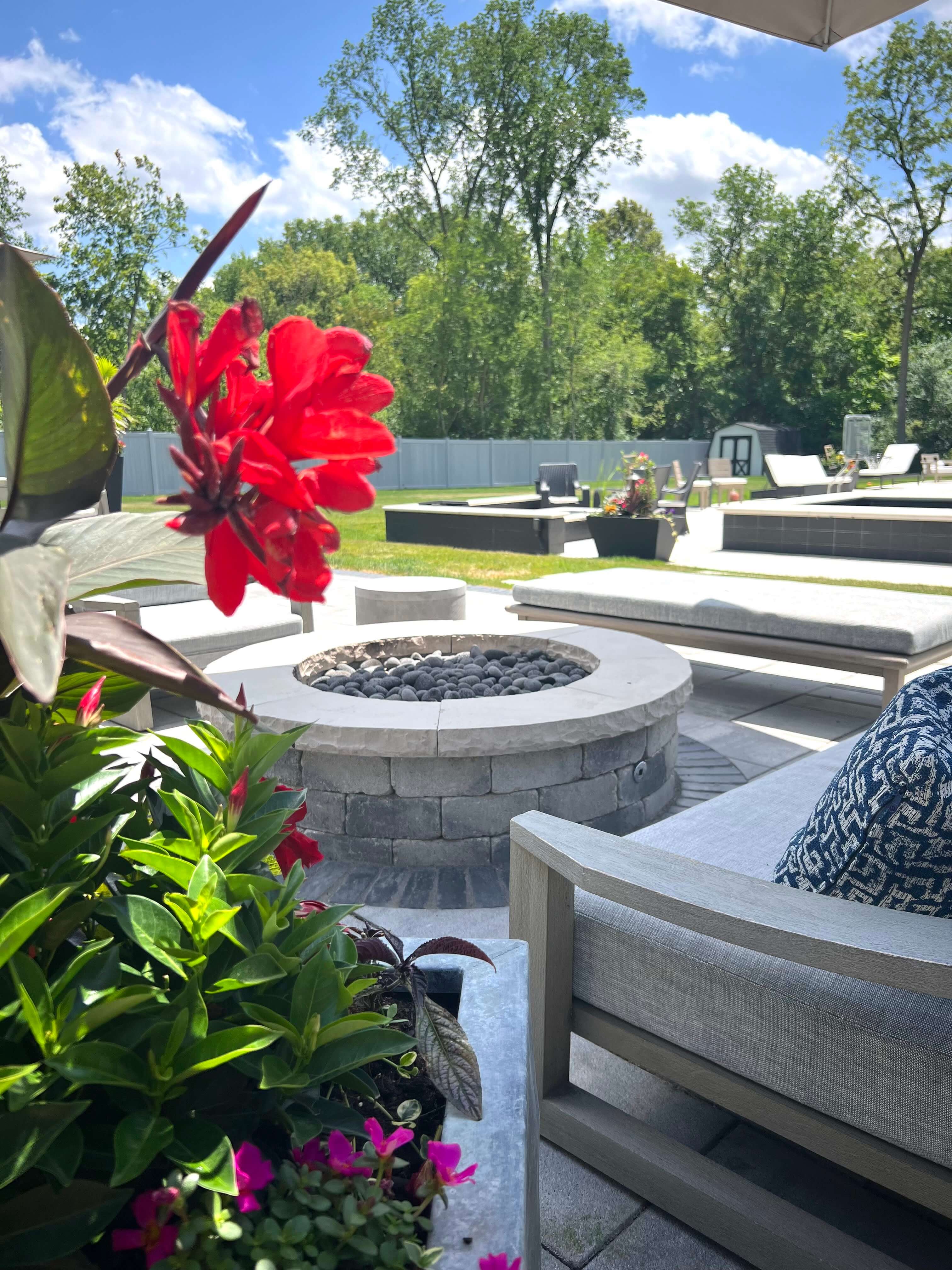 Outdoor patio with stone fire pit, cushioned seating, vibrant flowers, and lush green backyard trees.