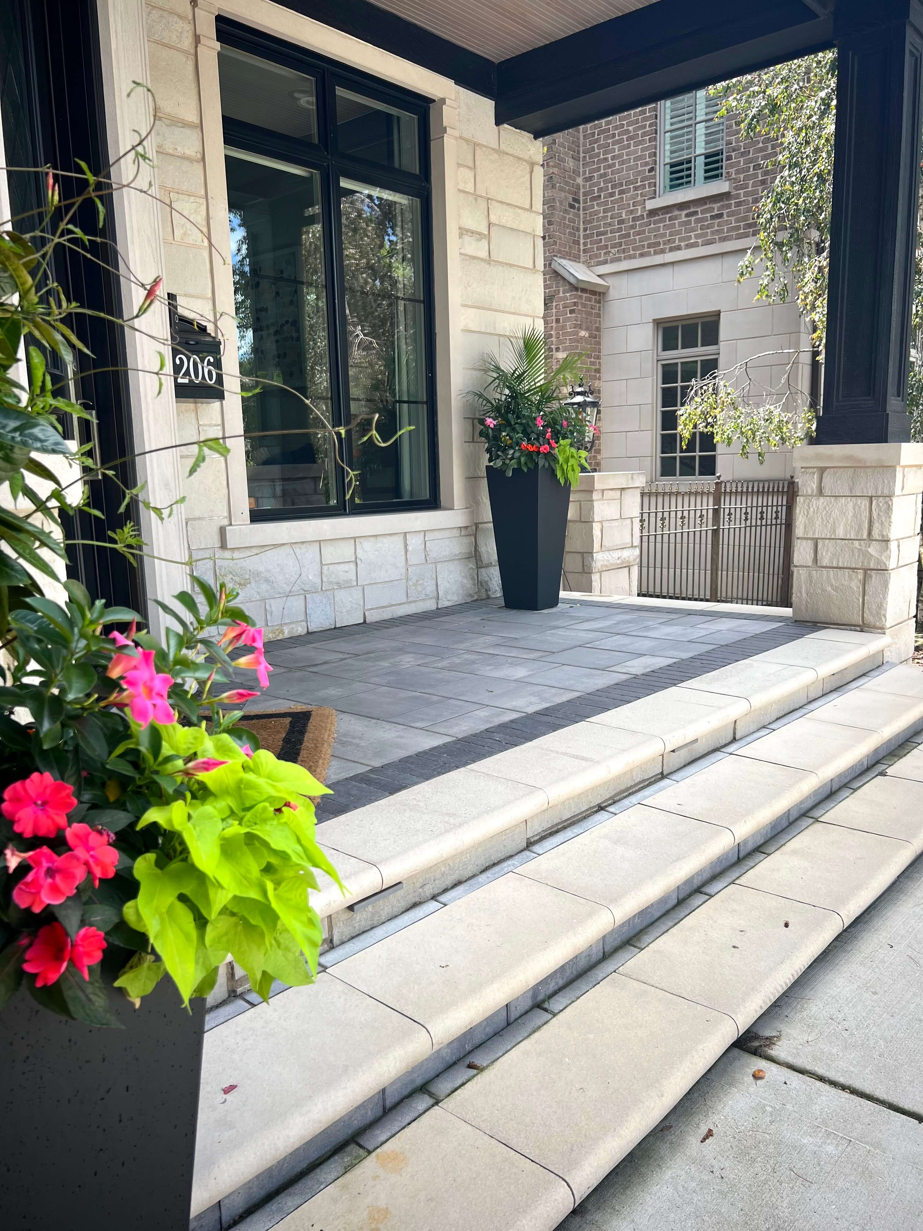 Front porch with stone steps, large black planters holding green and pink flowering plants.