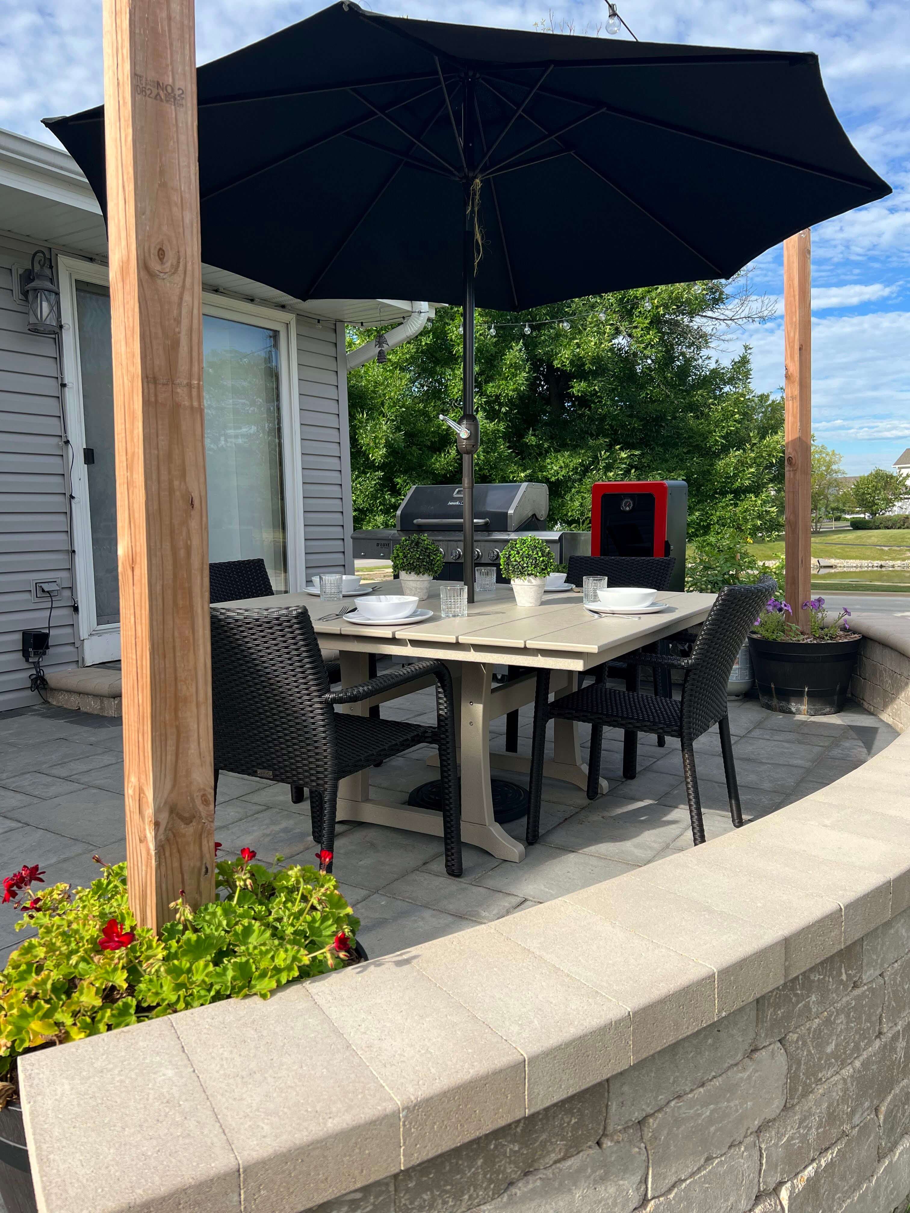 Outdoor patio with a dining table, four black wicker chairs, a large umbrella, and a grill in background.