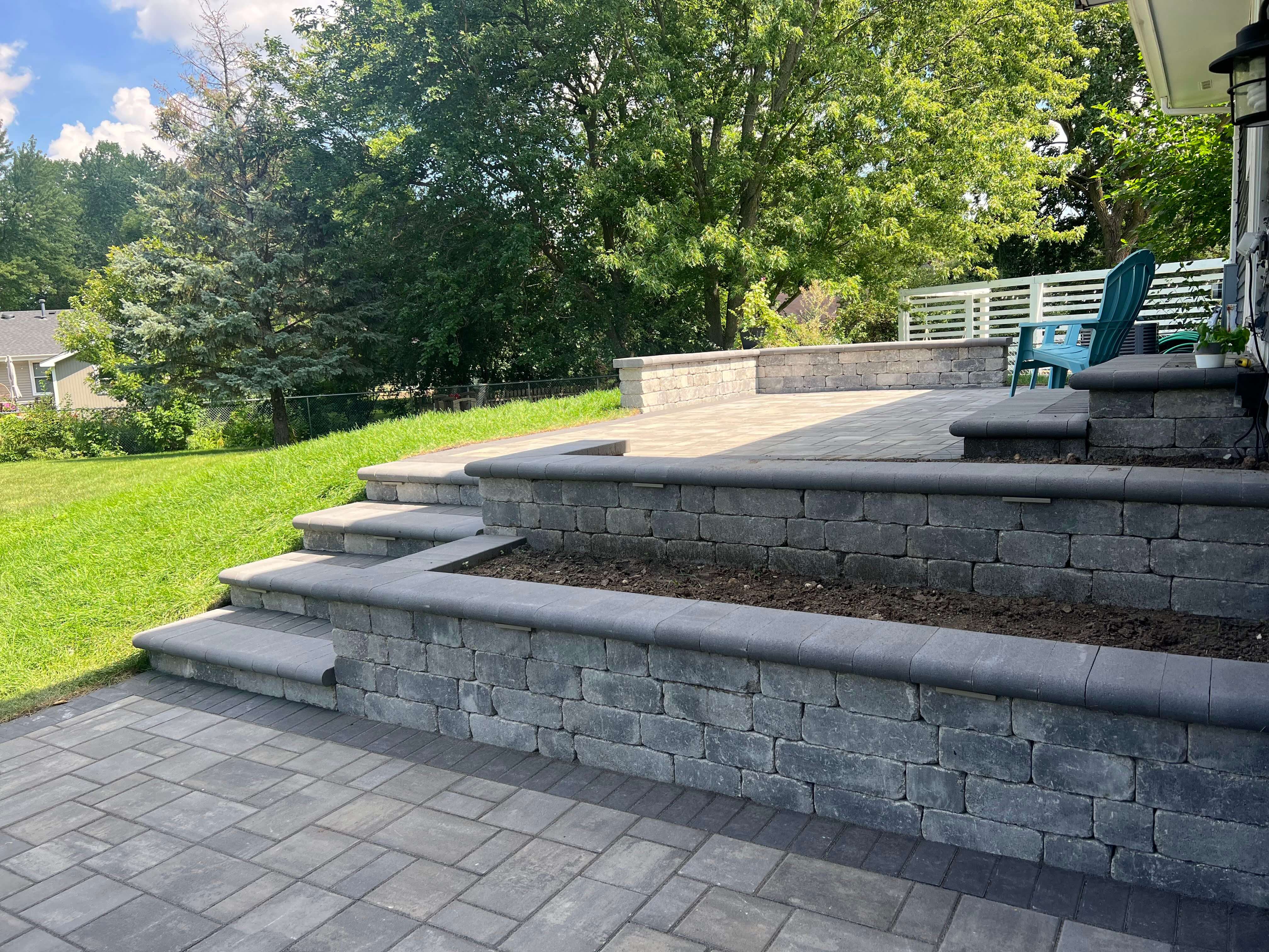 Square stone fire pit with glass rocks surrounded by gray Adirondack chairs on a beige stone patio.