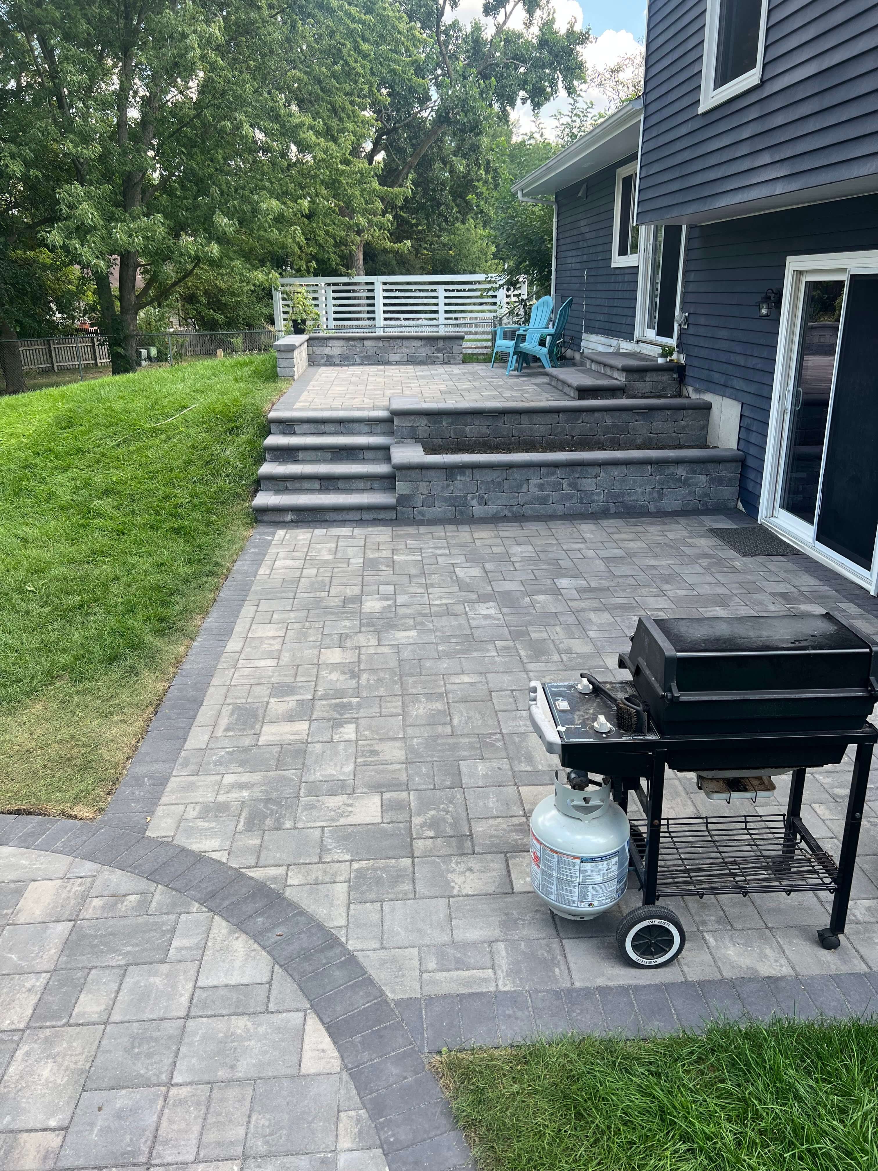 Raised gray stone patio with steps, retaining walls, blue chairs, black gas grill, and adjacent green lawn.